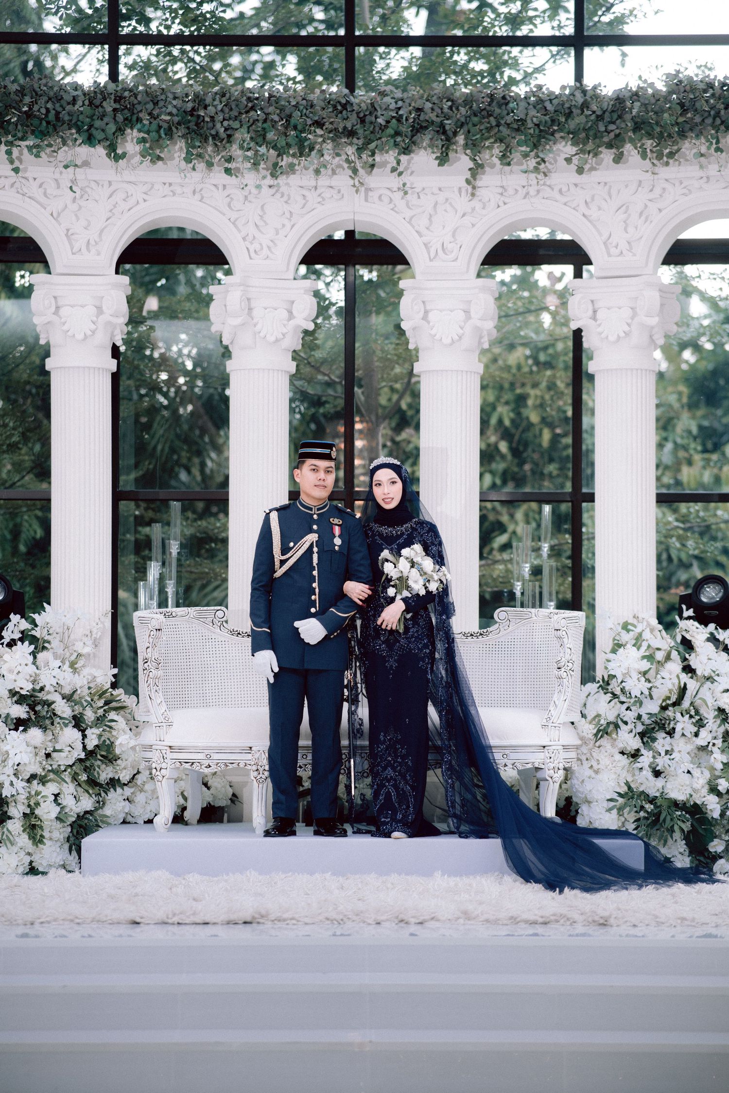 A couple in formal attire stands under white arched columns decorated with flowers for an elegant outdoor photo session.