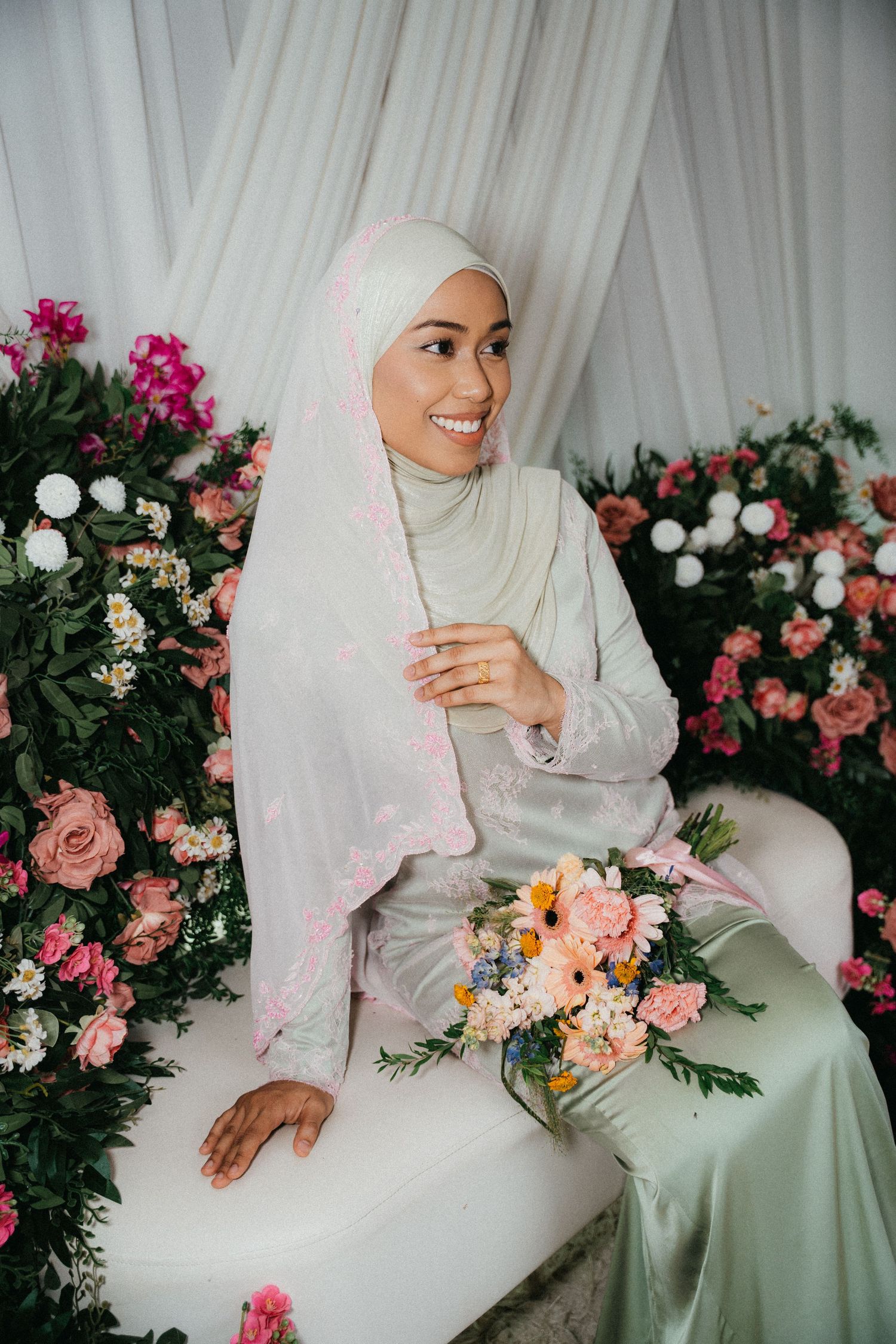 Beautiful bridal portrait with white hijab surrounded by pink and peach roses against white draped backdrop.