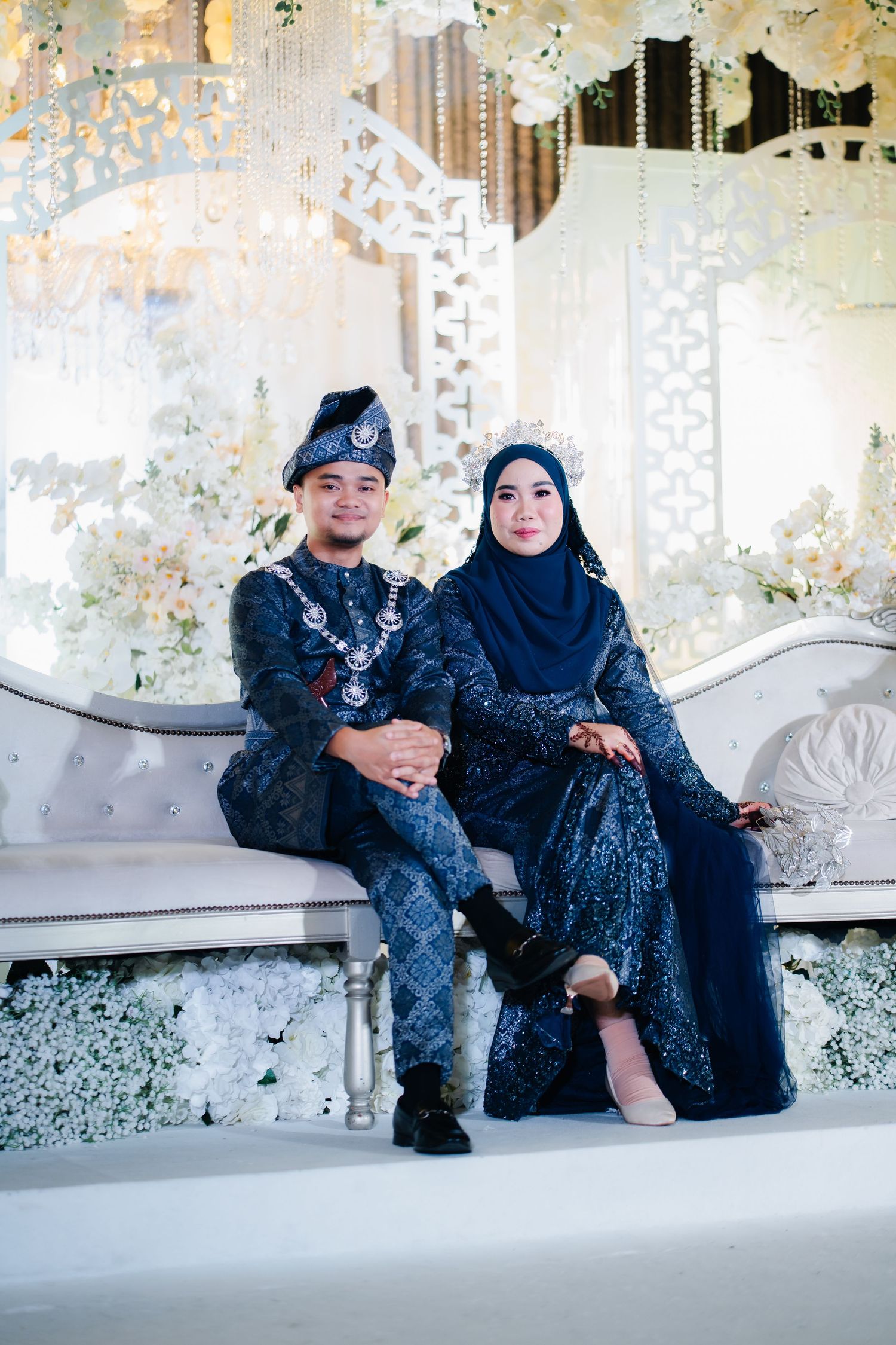 Two people in matching dark sequined outfits sit and pose together on an ornate white bench at an event venue.