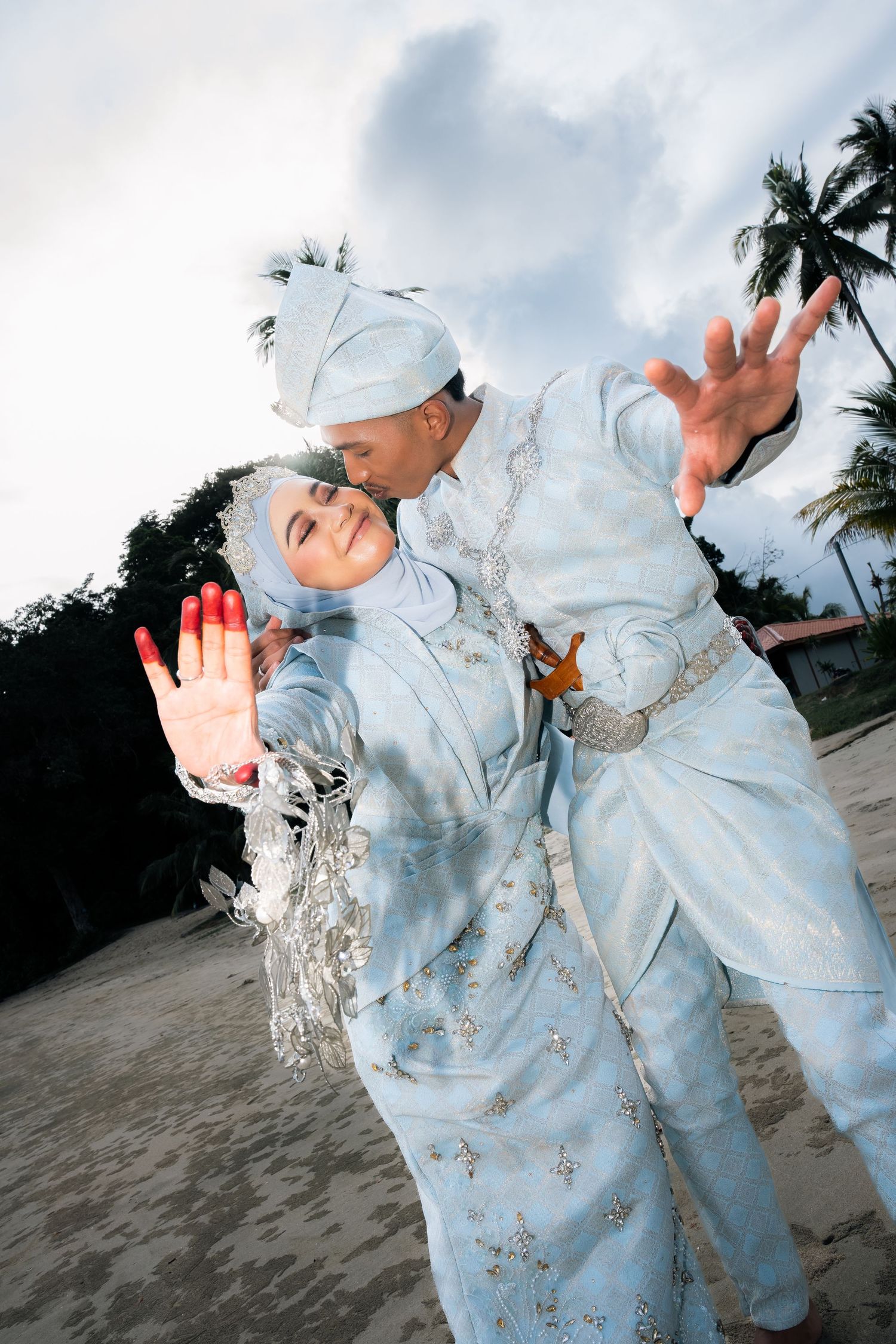 A romantic wedding couple in matching pale blue traditional outfits pose playfully on a beach with palm trees at sunset.