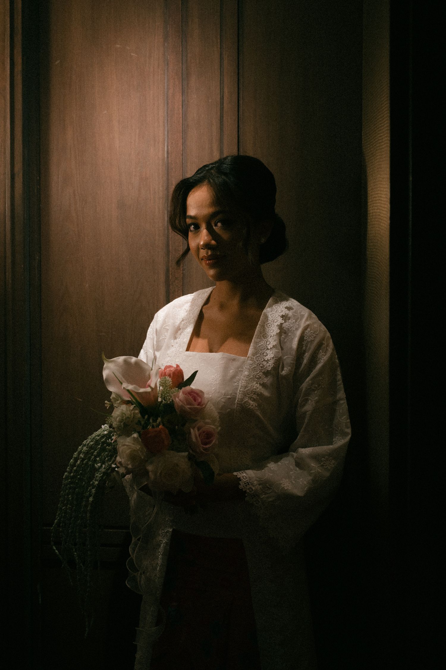 Moody portrait of a bride in a white dress holding pink roses against a dark wooden backdrop in vintage lighting.