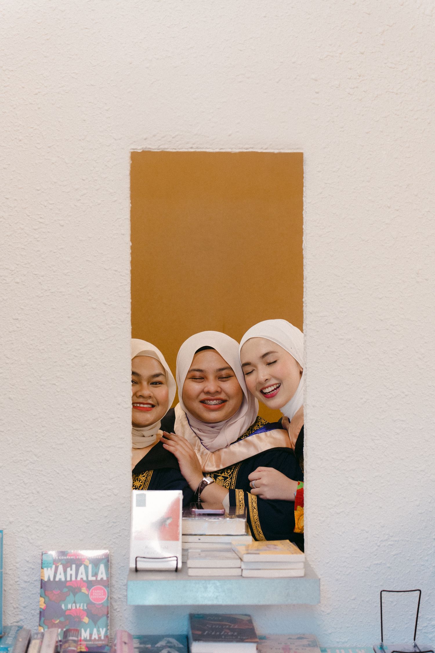 Three graduates posing together through opening in white wall with books visible.