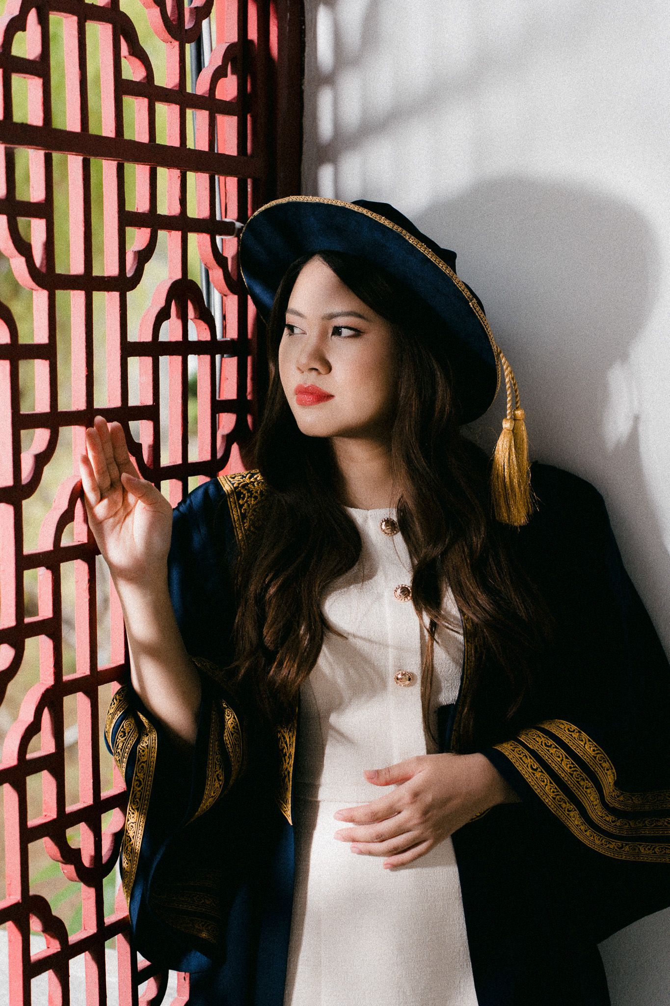 Young woman in graduation gown and cap with gold tassel poses elegantly beside ornate decorative window.