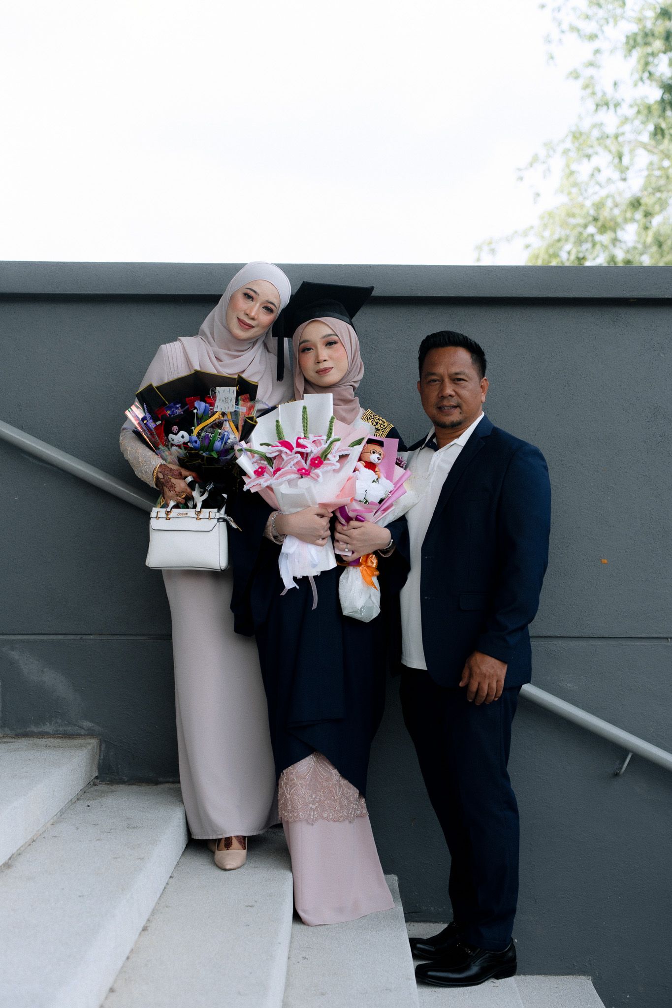 Muslim family celebrating graduation, graduate in cap holding flowers with parents on steps.