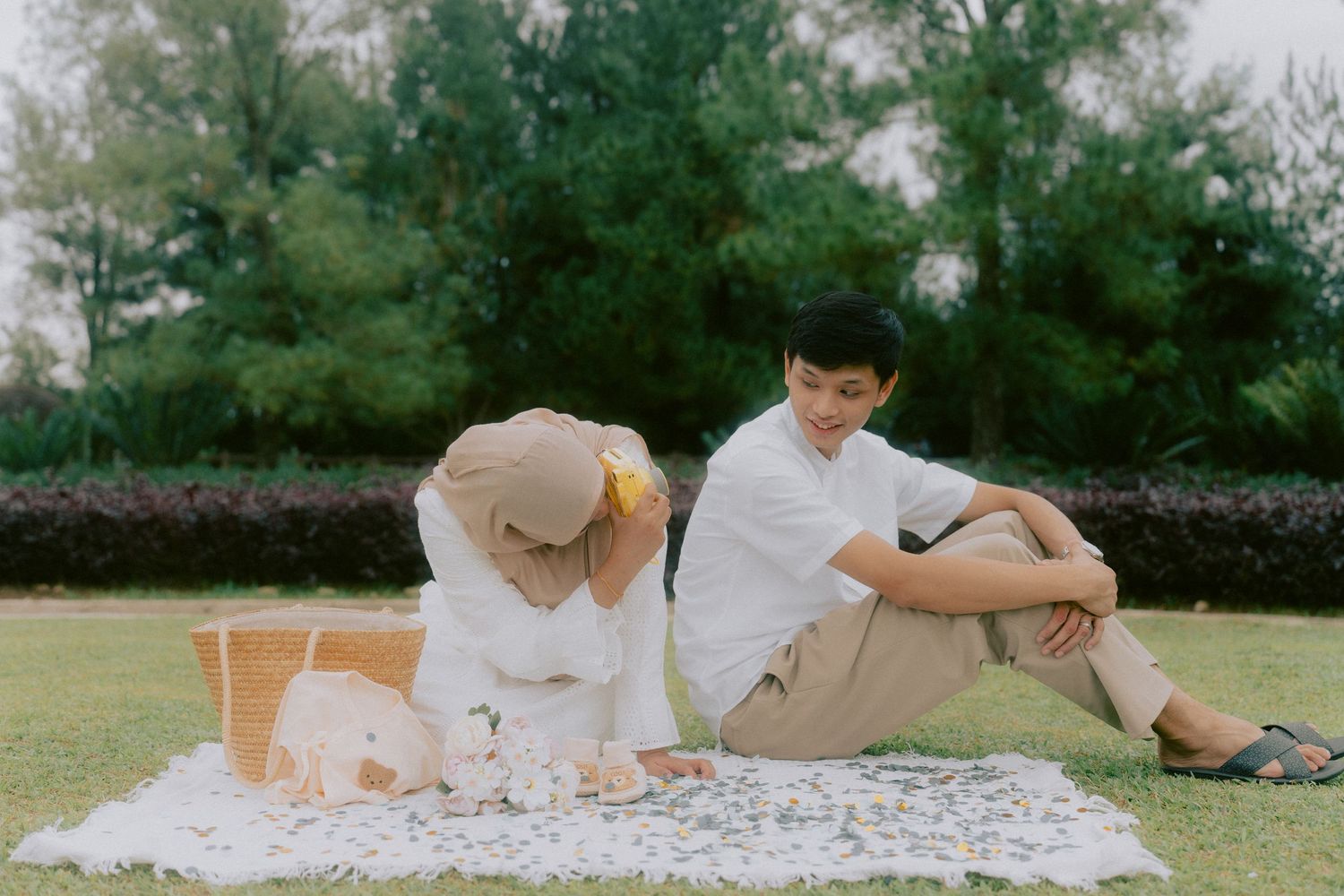 A couple relaxes on a picnic blanket in a garden setting with trees in the background.