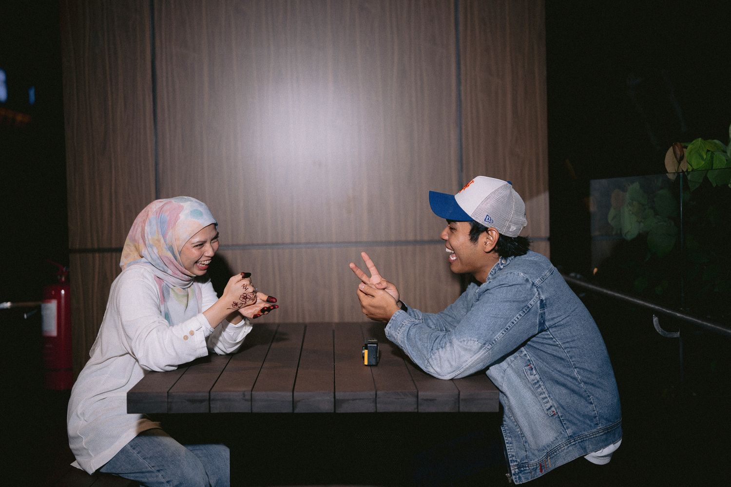 Two people in casual wear sit across a table, engaged in animated conversation in a dimly lit room.