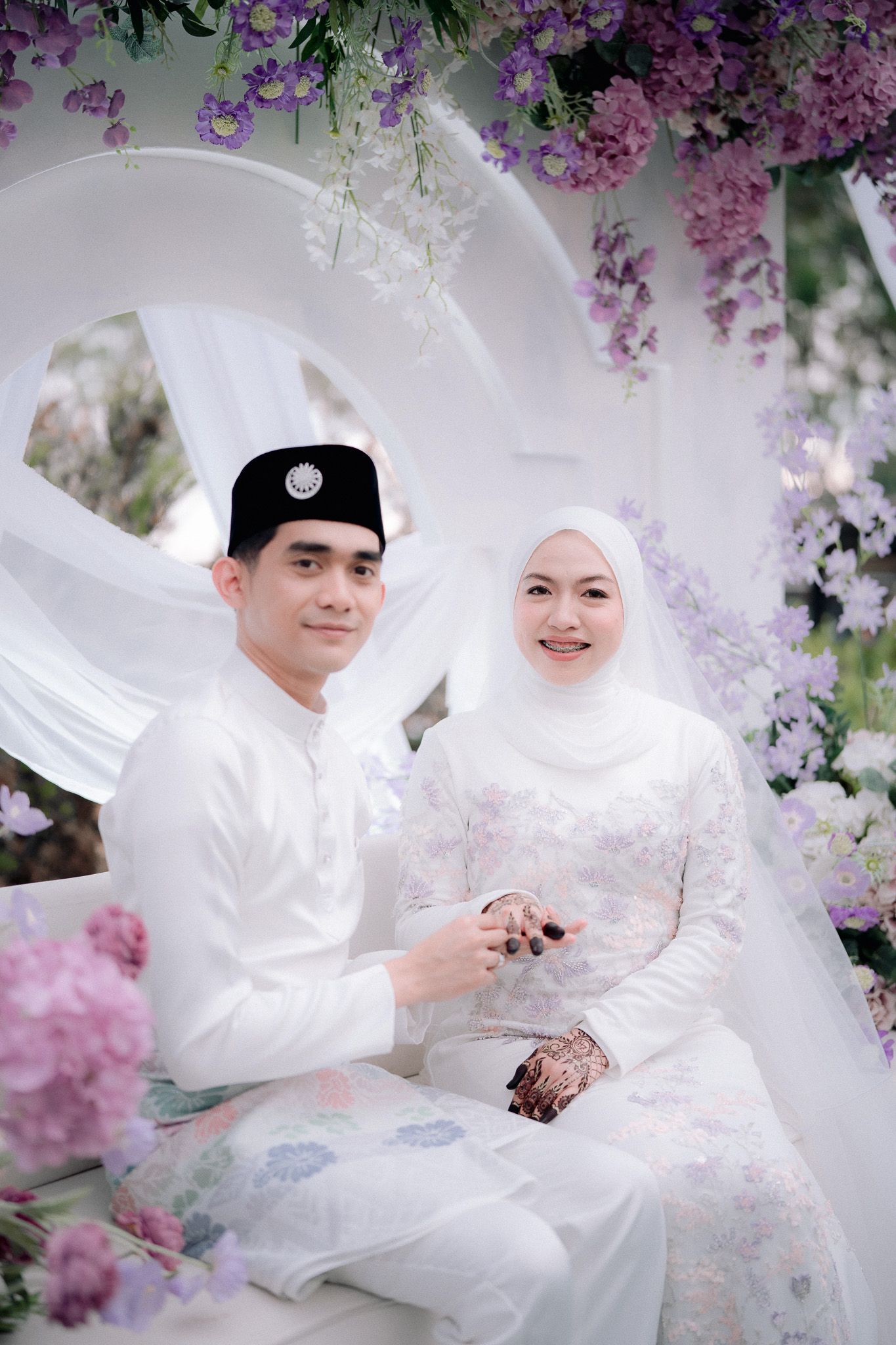 Malay couple in white traditional attire sitting together at a floral wedding ceremony with purple and white flowers.
