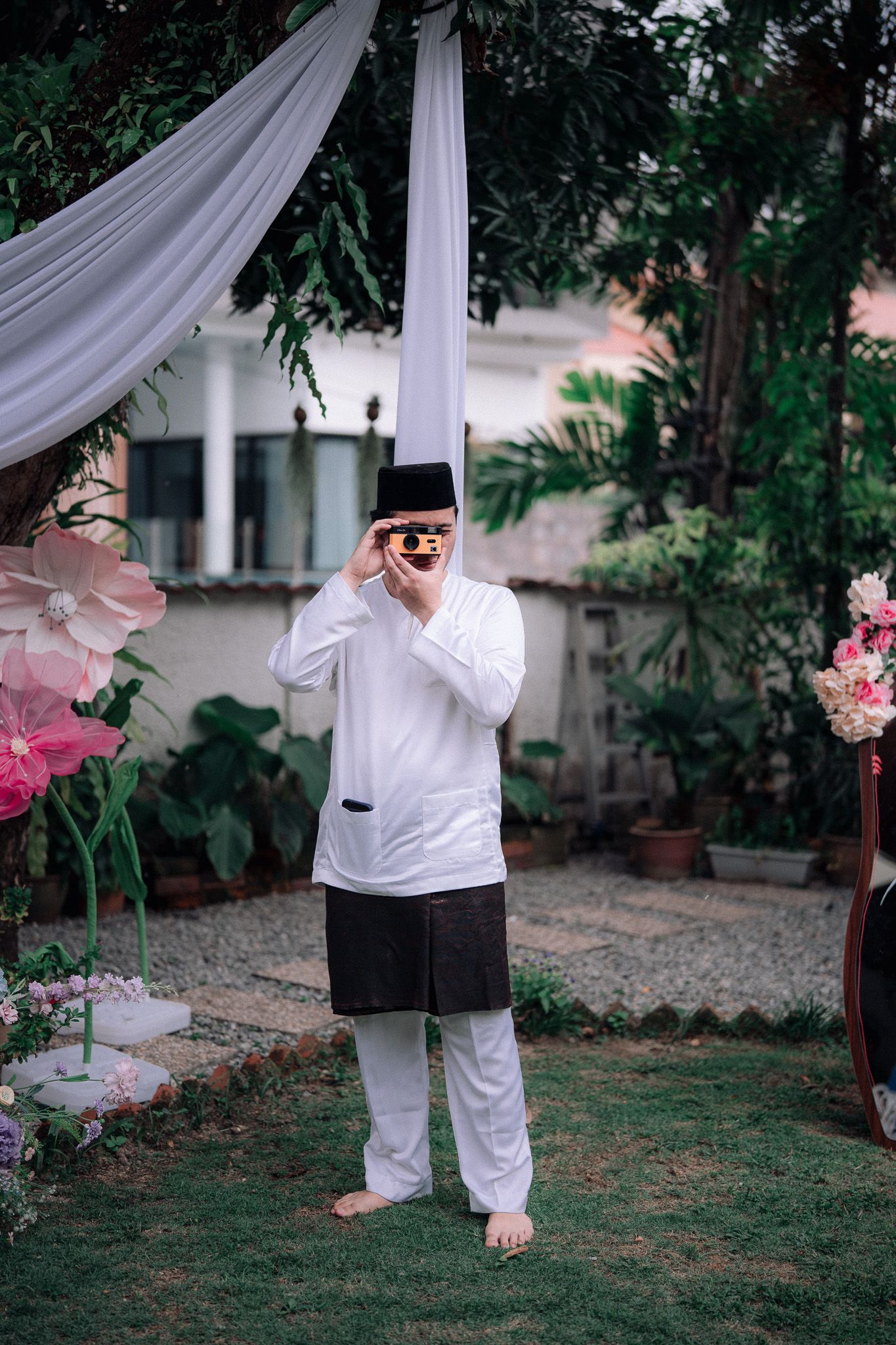 Man in traditional white Baju Melayu and black songkok hat photographing outdoors at a decorated garden event.