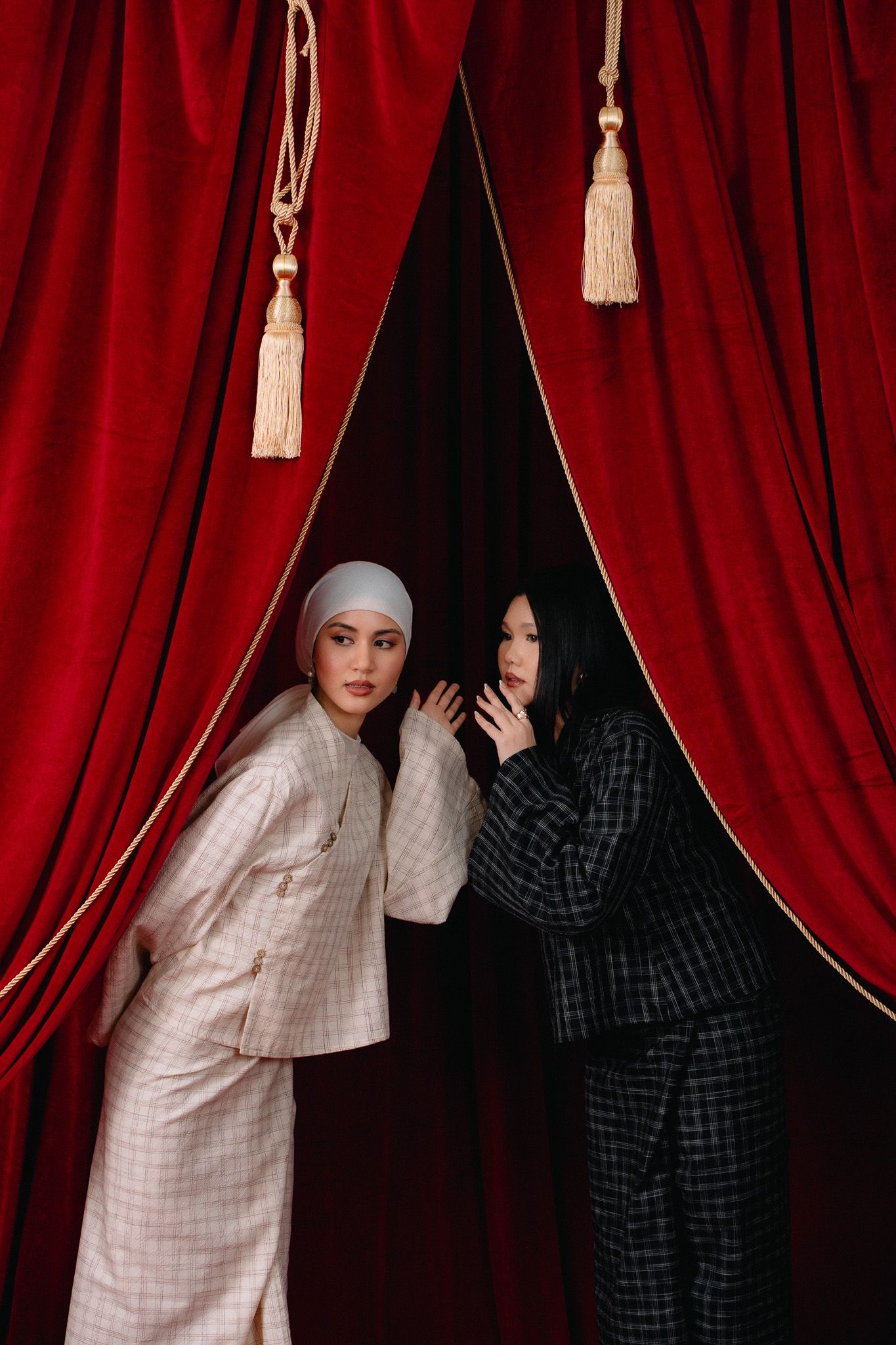 Two women in plaid suits pose between red velvet curtains with tassels, one wearing a white hijab.