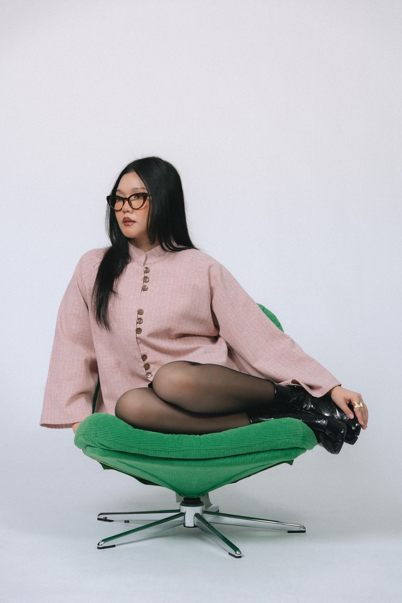 Woman in pink sweater and glasses sits cross-legged on a green retro chair against a white background.