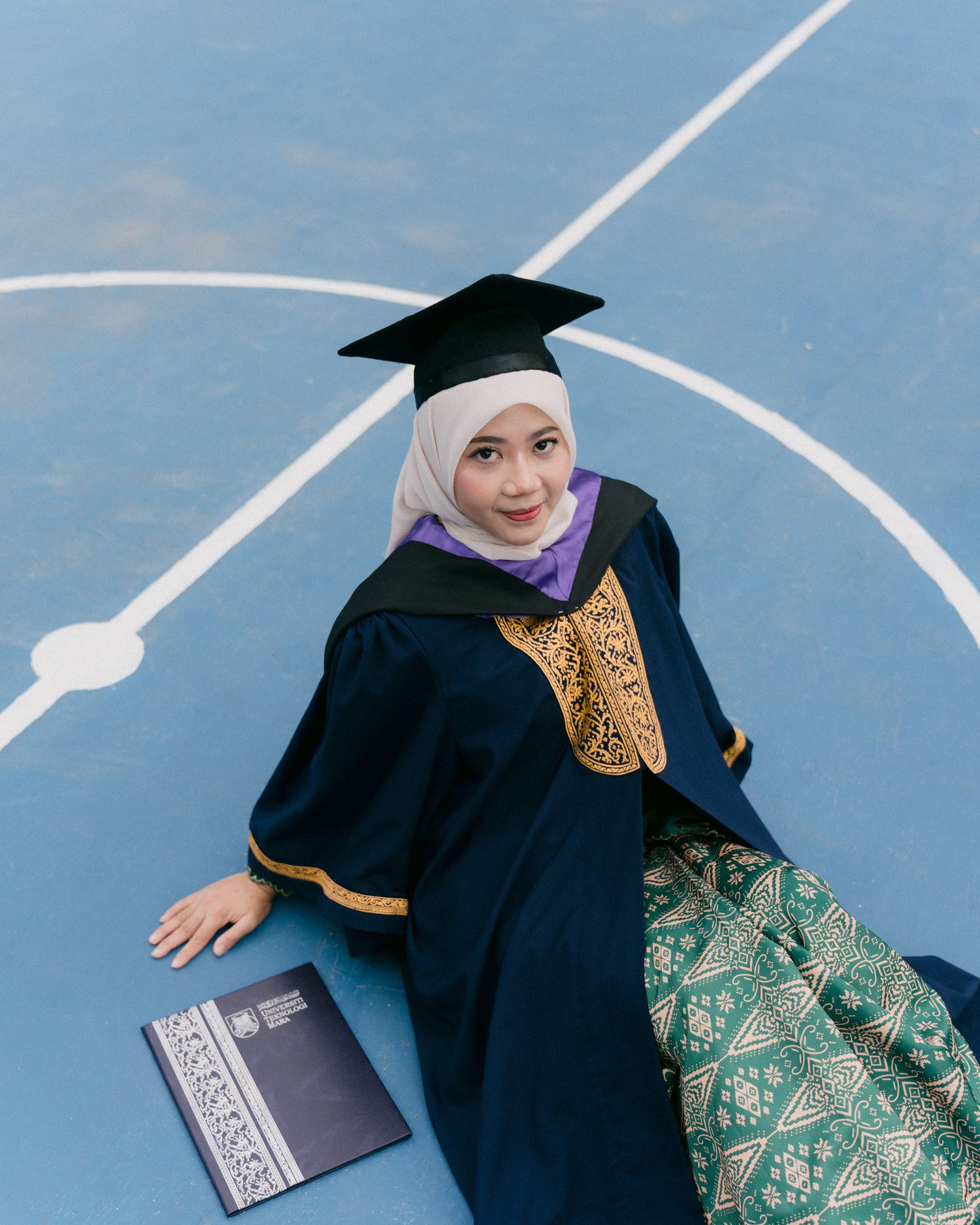Graduate in black cap and gown with green traditional dress celebrating achievement on blue background.