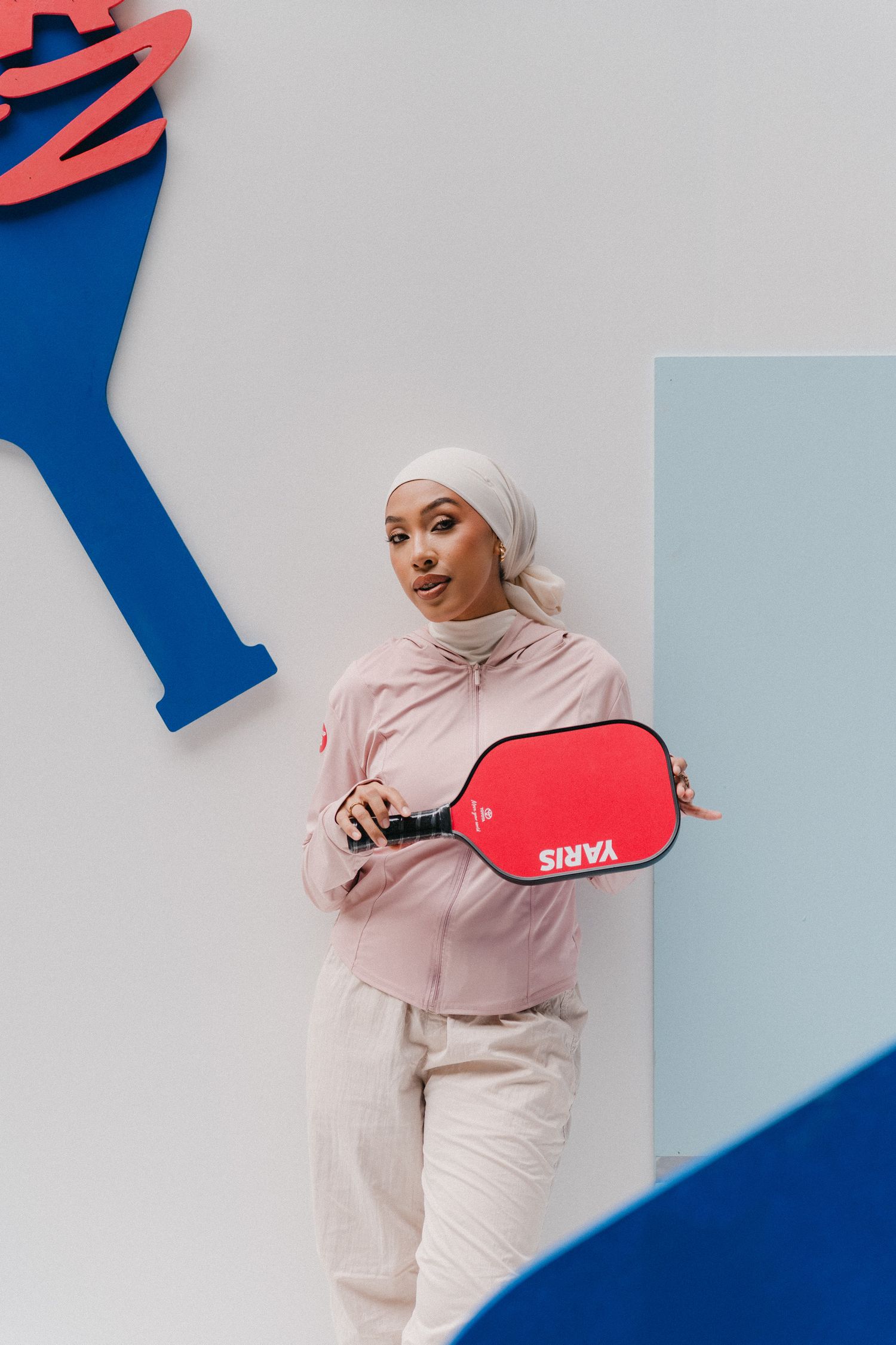 Athletic model in light hijab and sportswear poses against blue and white backdrop for fashion photoshoot.