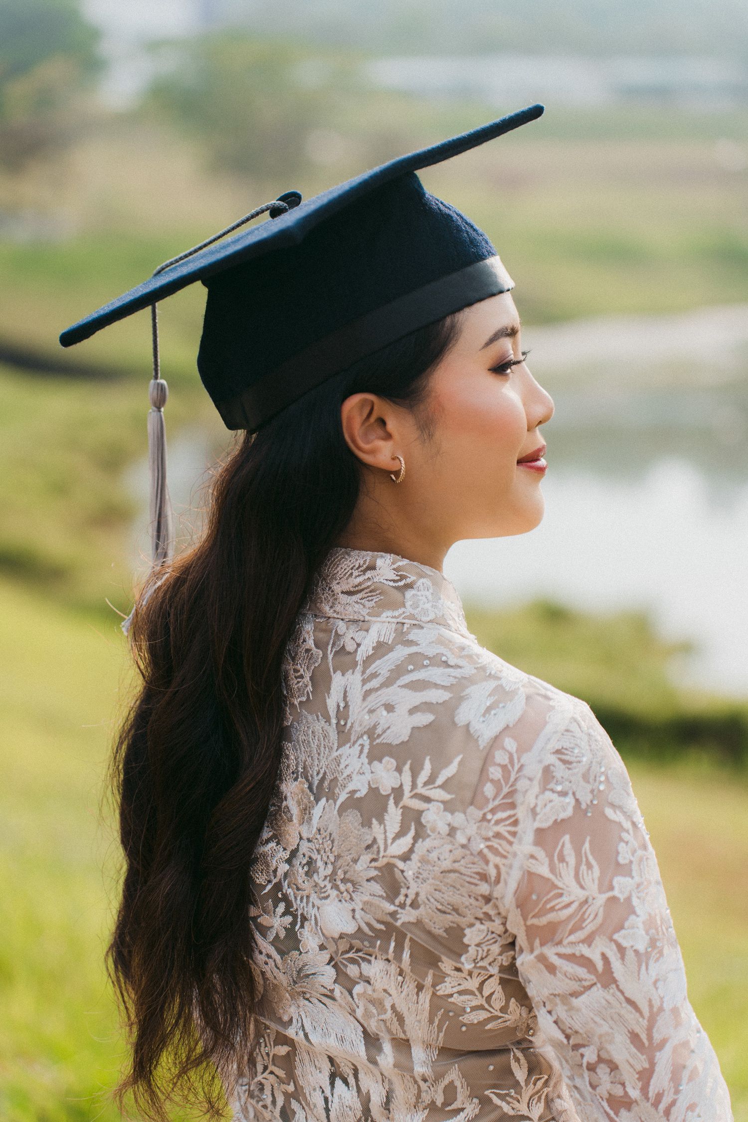 Graduate in white lace dress and mortarboard cap poses against blurred natural landscape background.