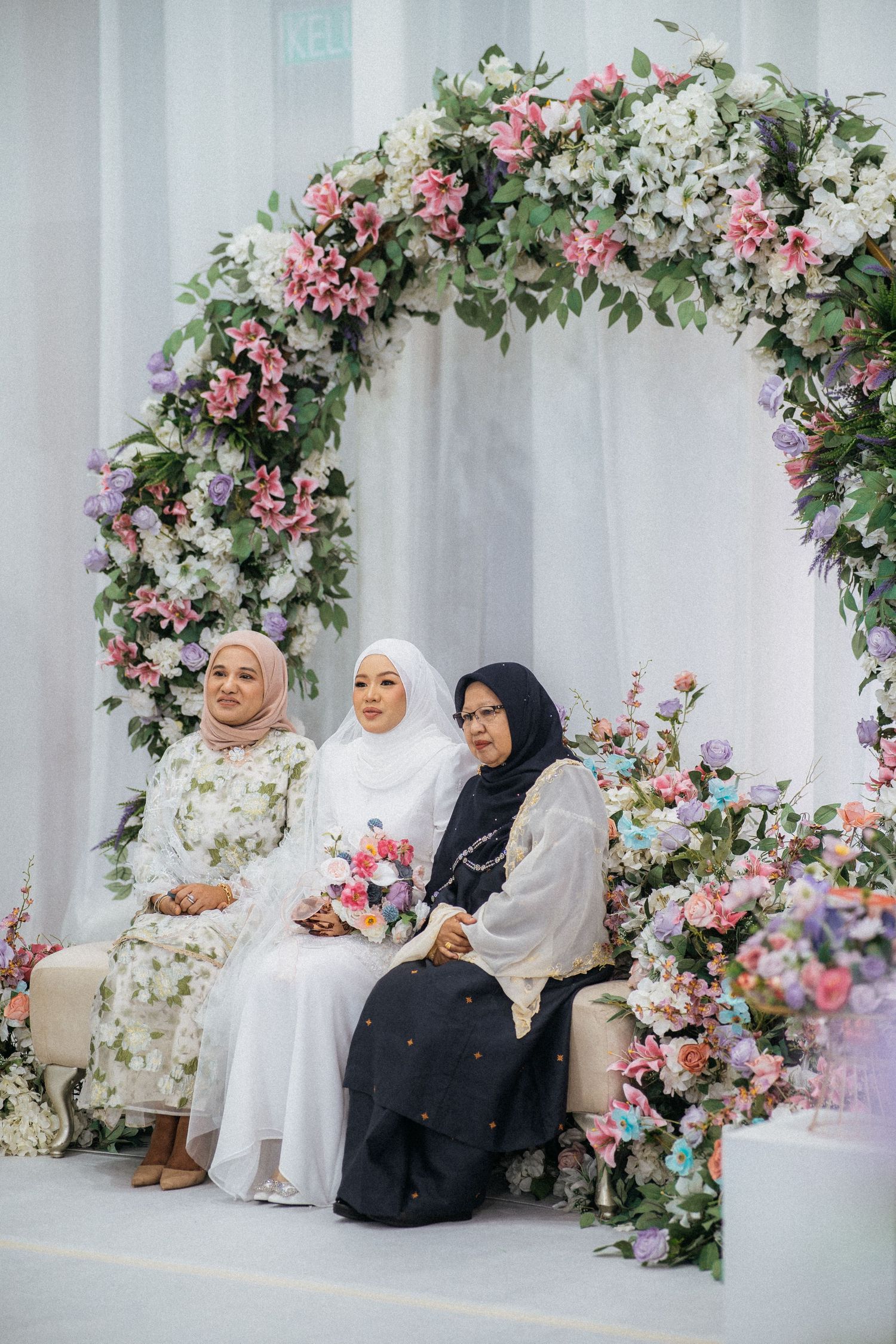 Beautiful floral arch with pink and white flowers decorates a traditional celebration setting with guests in hijabs.