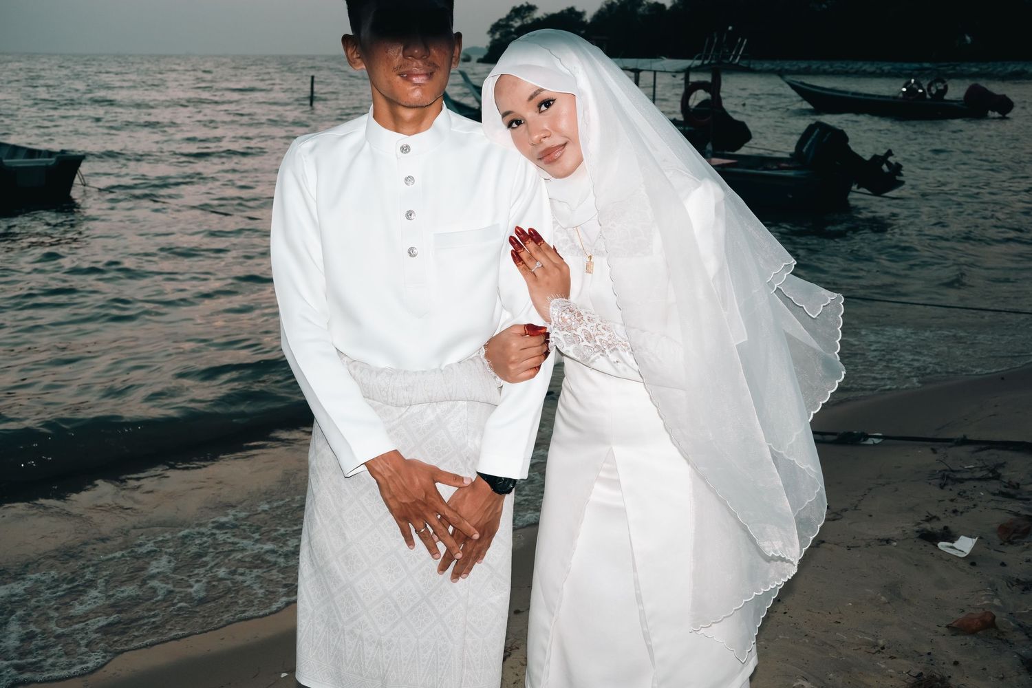 Newlywed couple in white attire strolling along a beach at sunset.