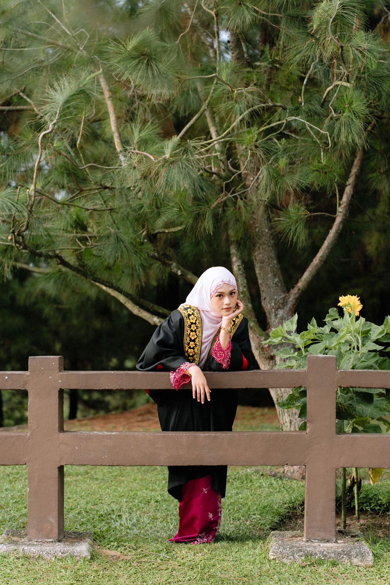 A Muslim woman in black abaya and hijab leans on a wooden fence in a lush green park with trees and a sunflower.