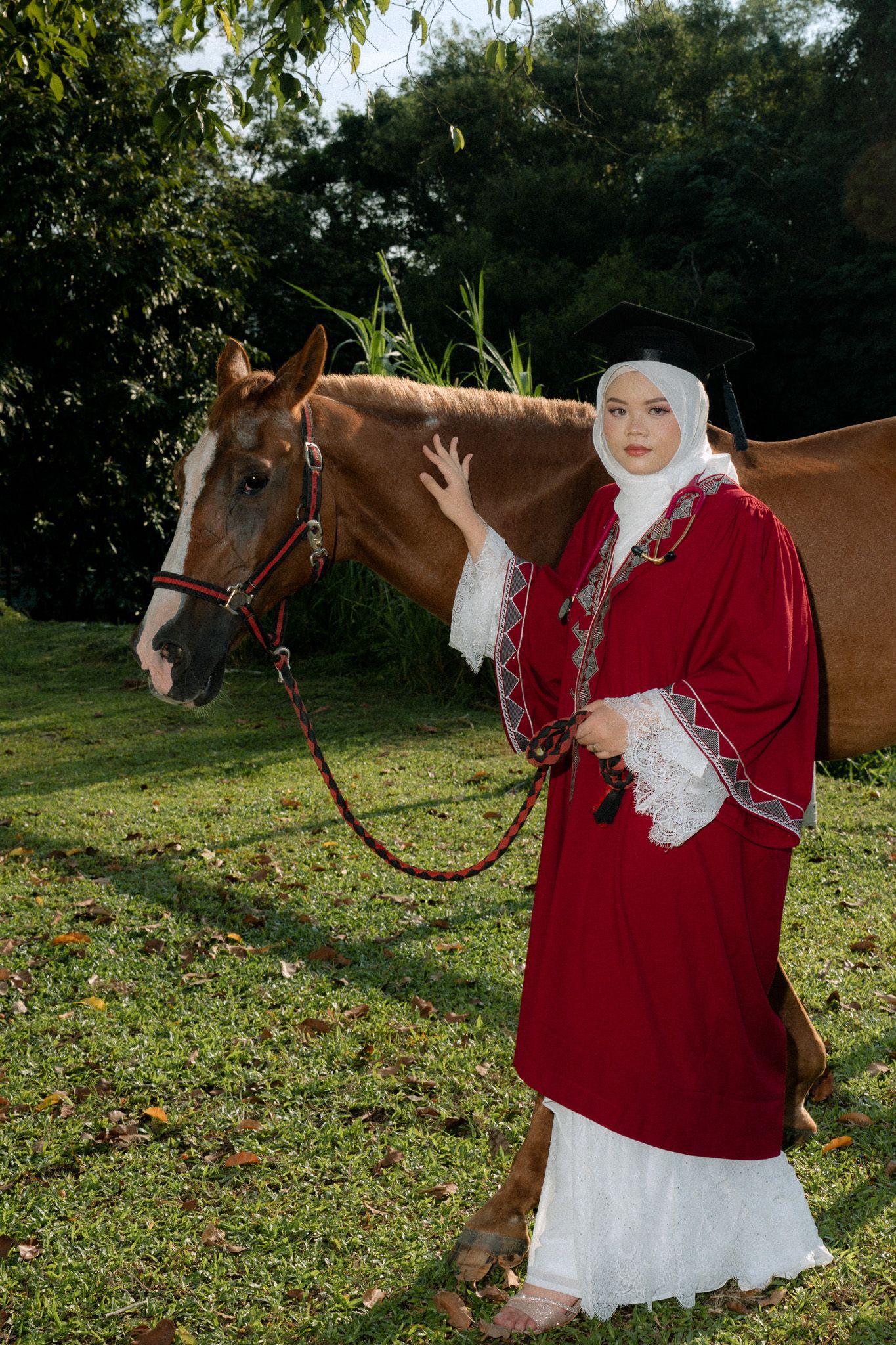 Muslim woman in red graduation gown and hijab poses with a brown horse on green grass outdoors.