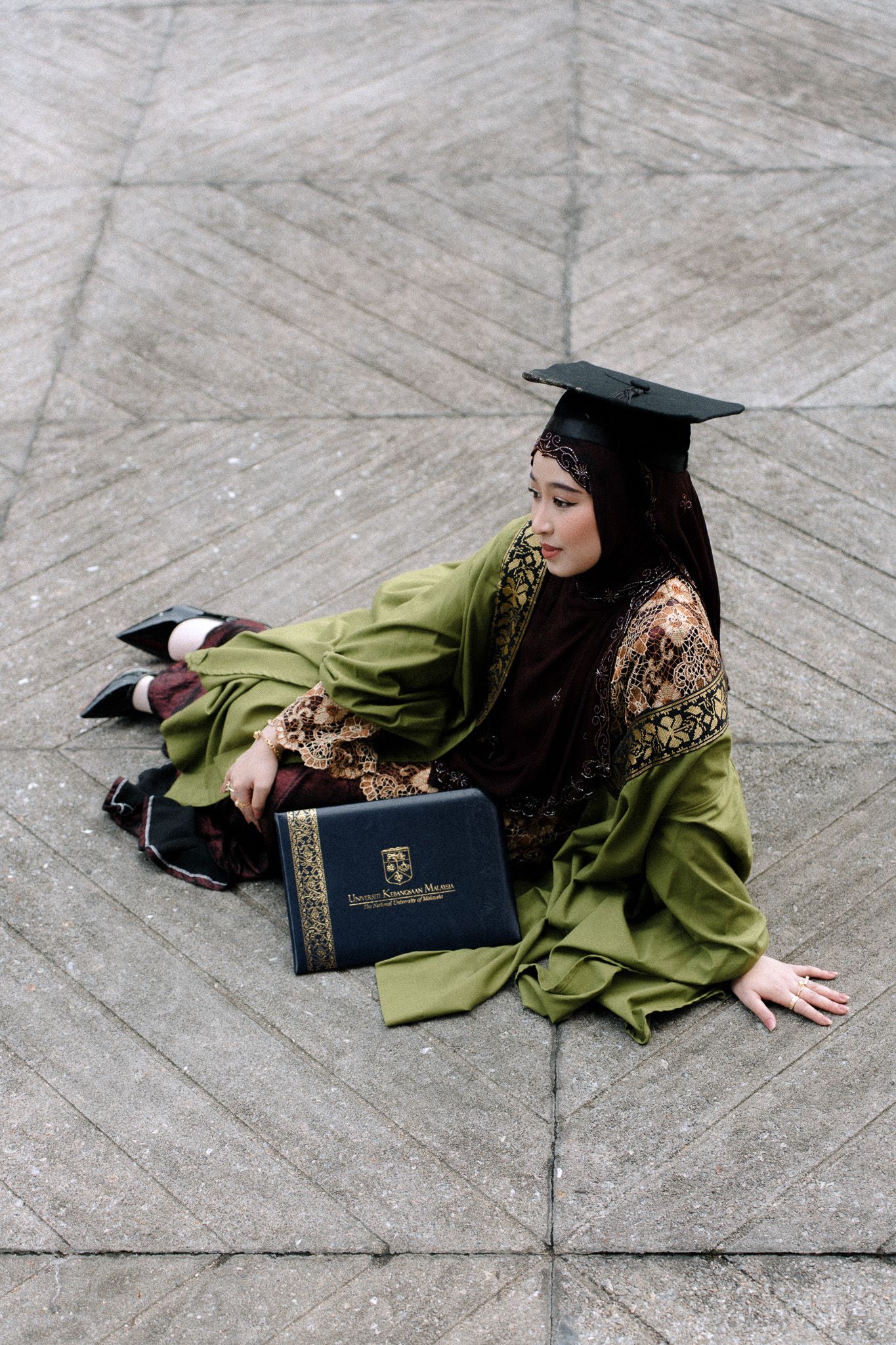 Muslim woman in green graduation robe and mortarboard sits on ground holding diploma, celebrating academic achievement.