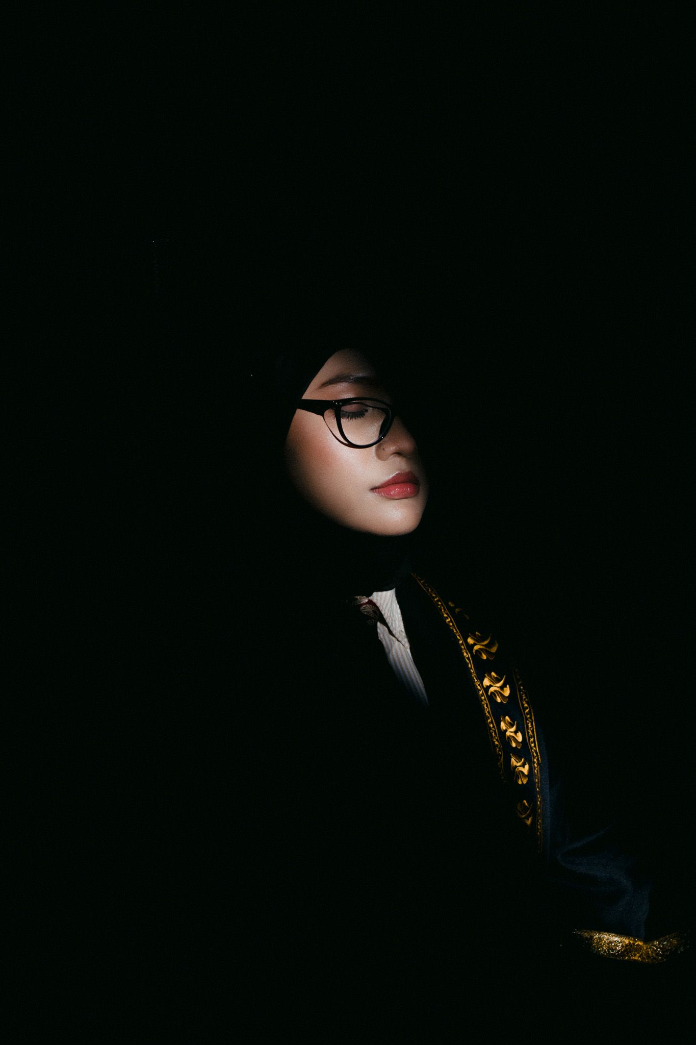 A young woman wearing hijab and glasses poses dramatically in low-key lighting against a dark background.