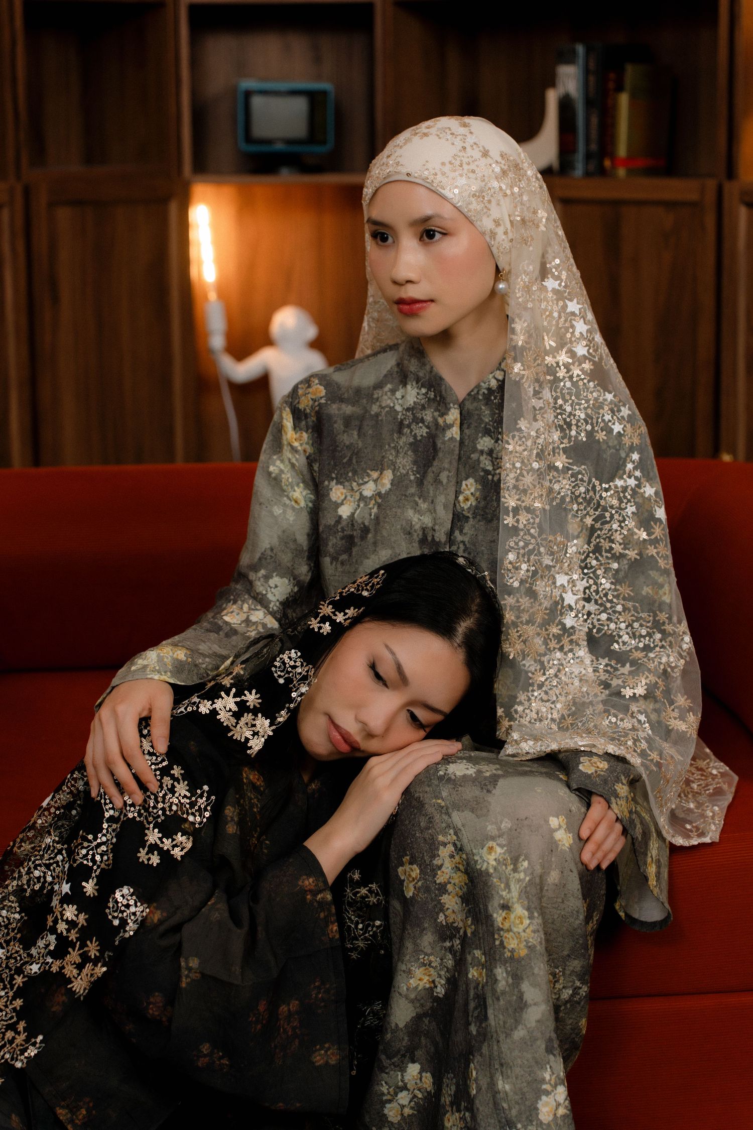 Two women in traditional floral dresses sit on a red couch, one resting her head on the other's lap.