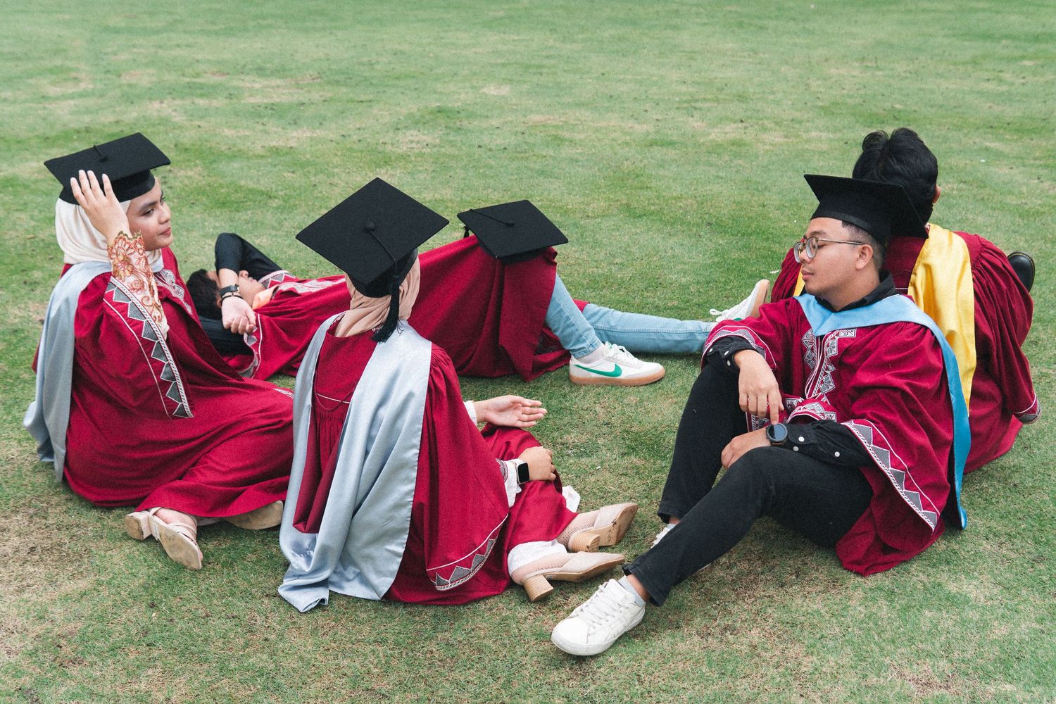Graduates in red gowns relax on grass during graduation photo session.