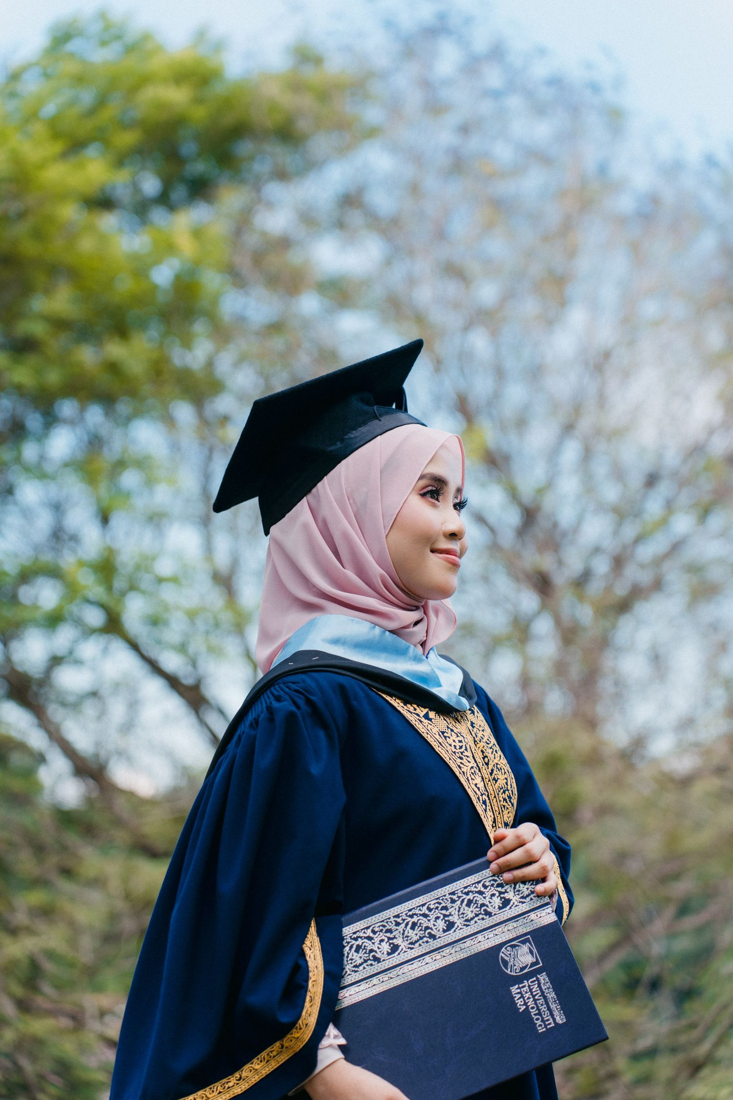 Graduate in navy blue academic regalia and pink hijab poses outdoors with diploma during commencement ceremony.