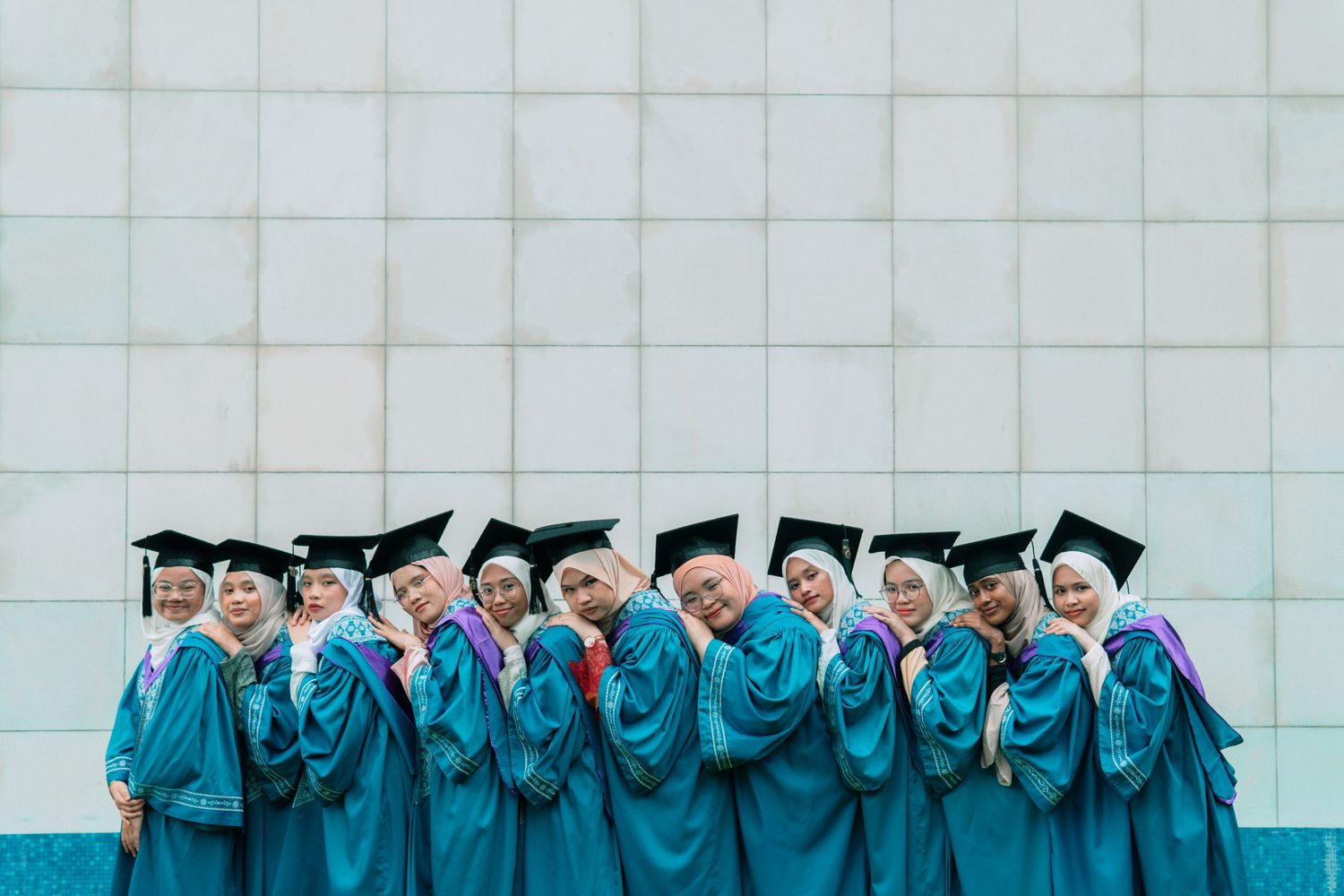 Row of graduates in teal gowns standing against white wall for group photo.