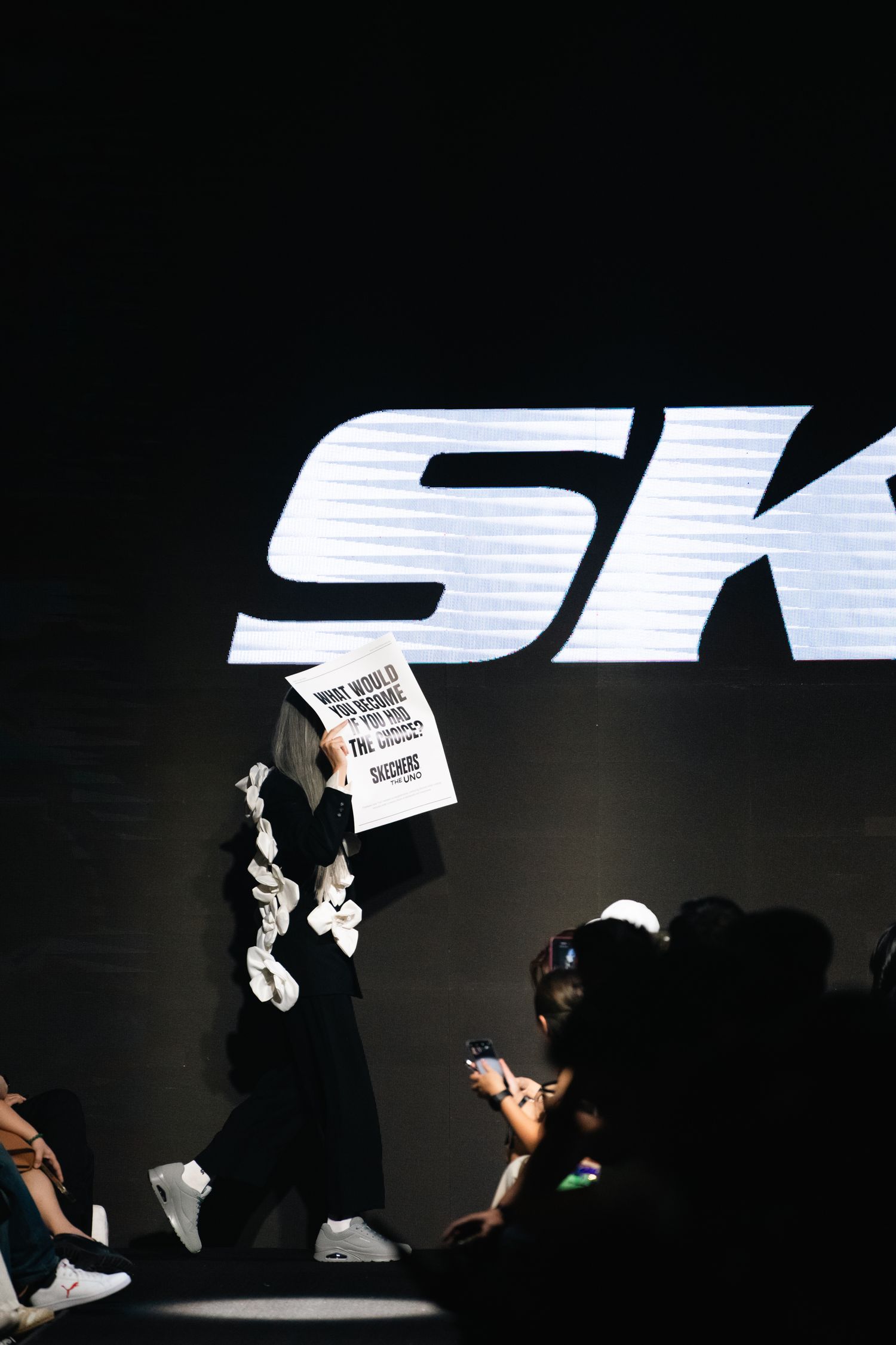 A dramatic spotlight illuminates the Skechers logo on a dark stage during a fashion show.