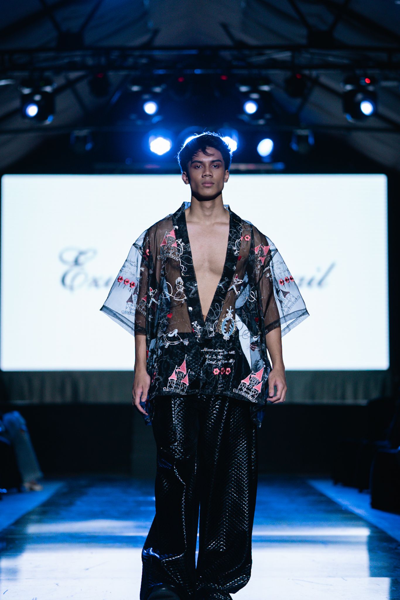 Model wearing floral sheer robe and dark pants walks runway under blue lighting.