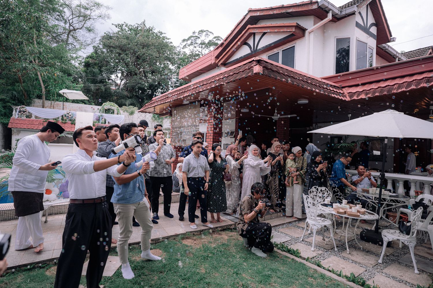 Guests celebrate outdoors at a festive garden party with confetti near a charming house with a red roof.