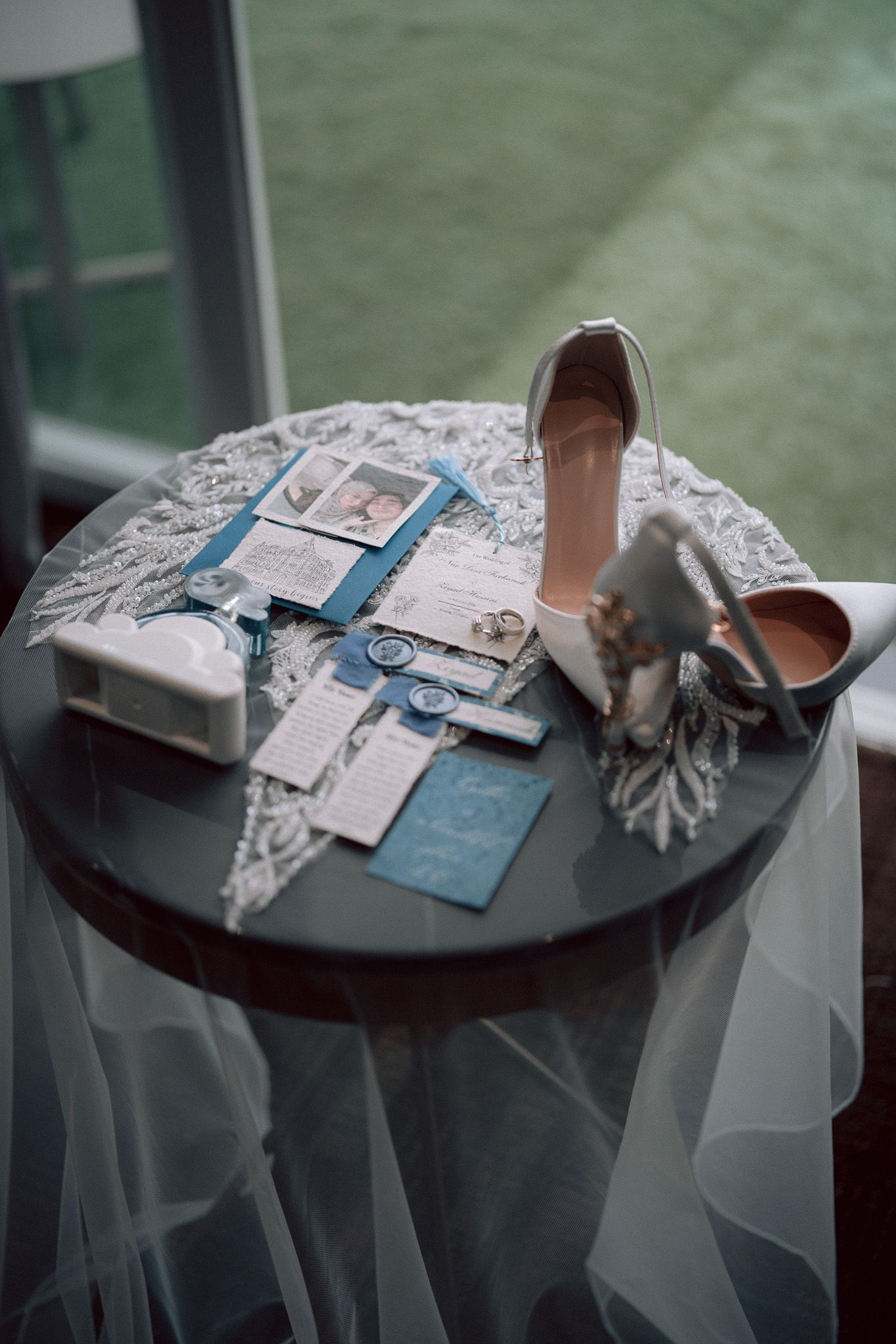Elegant wedding flat lay with white heels, blue invitations, rings, and lace veil on a round table.