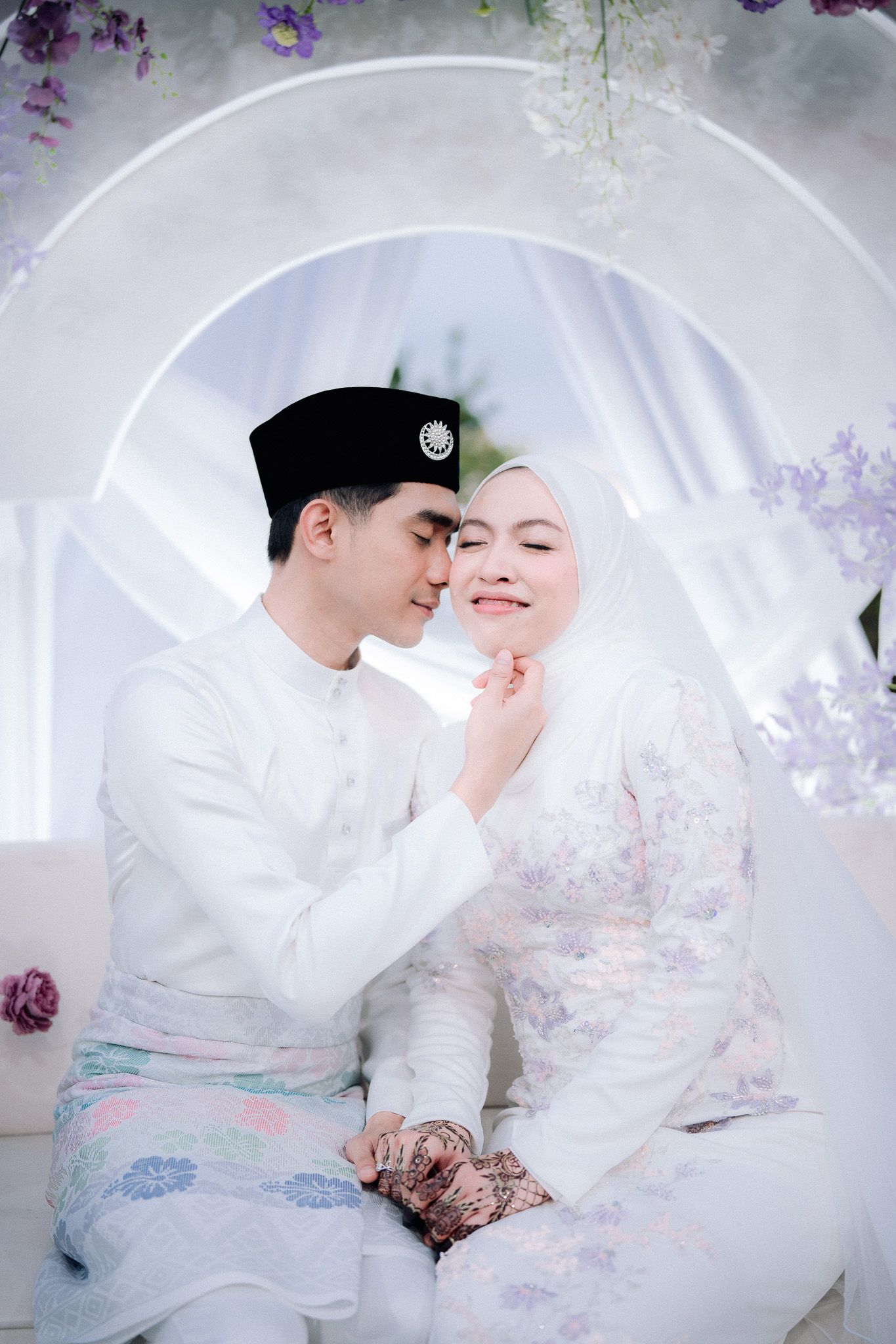 Malay couple in traditional white wedding attire sitting together under a floral arch, groom in songkok touching bride's face.