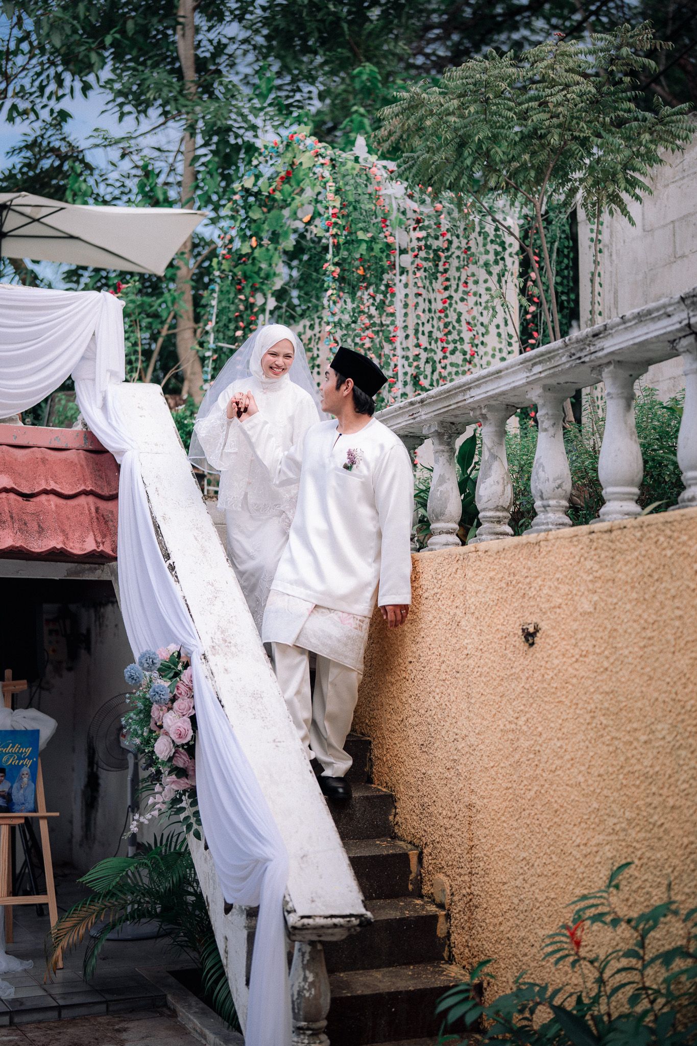 Muslim couple in white wedding attire walking up decorated stairs with floral arrangements and greenery backdrop.