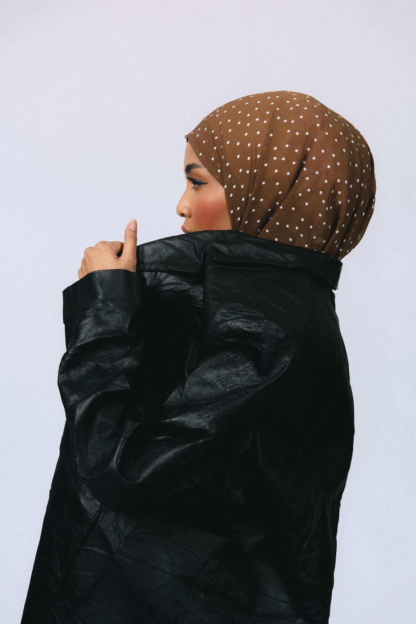 Woman in black leather jacket and brown polka dot hijab posing sideways against a light gray background.