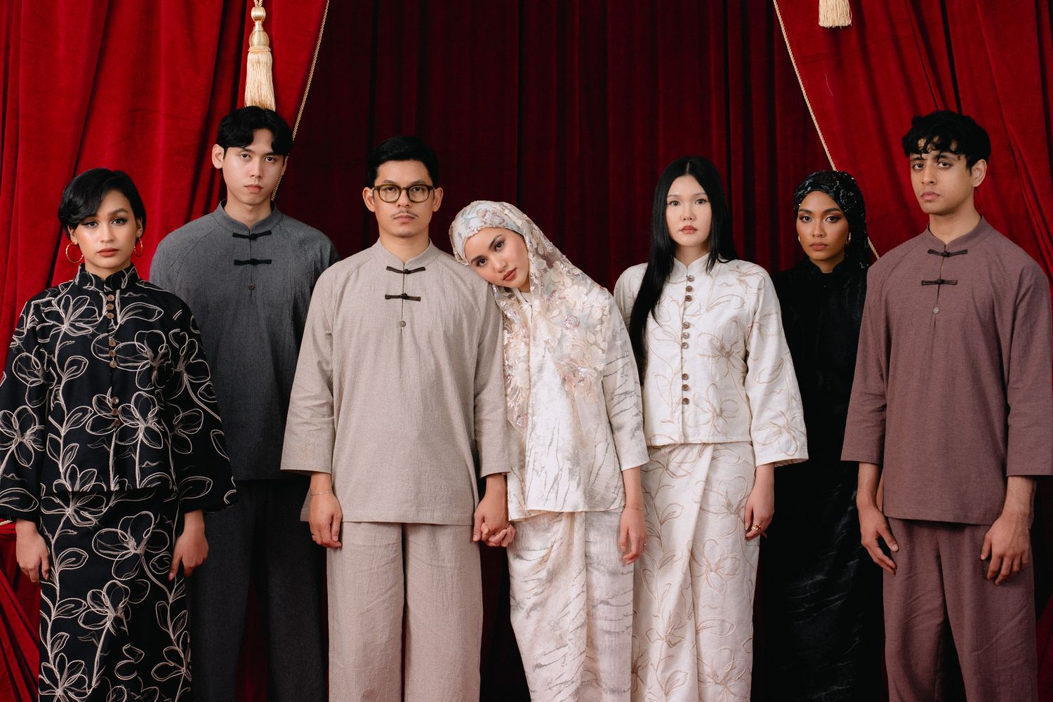Seven people in elegant traditional Asian attire pose together against red curtains in a formal group portrait.