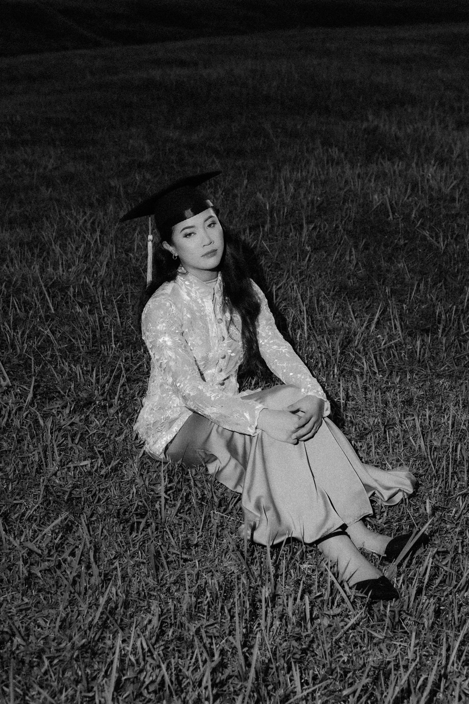 A figure in a white lace dress sits gracefully in a grassy field captured in black and white photography.