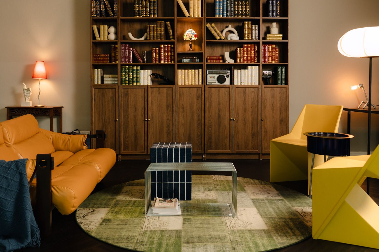 Modern library lounge with orange leather sofa, yellow armchair, wood bookshelf, and glass coffee table on green rug.