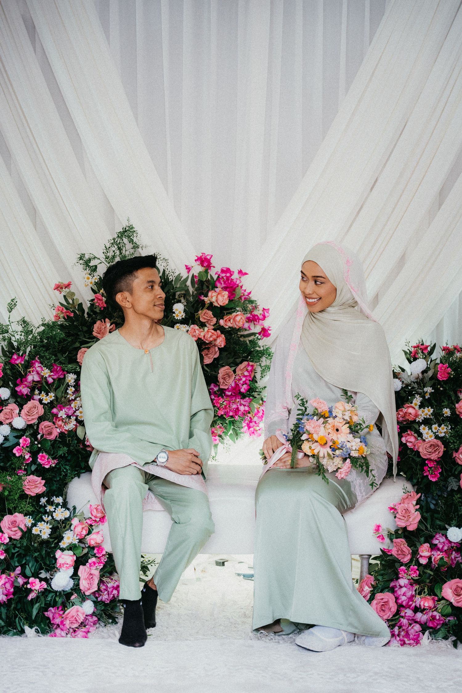 A couple in mint green outfits sits together on white cushions surrounded by pink floral arrangements and draping.