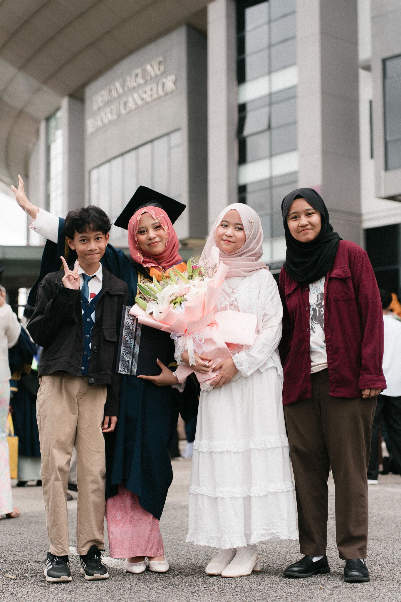 Muslim family celebrating graduation outdoors, graduate in cap and gown holding flowers with siblings posing together.