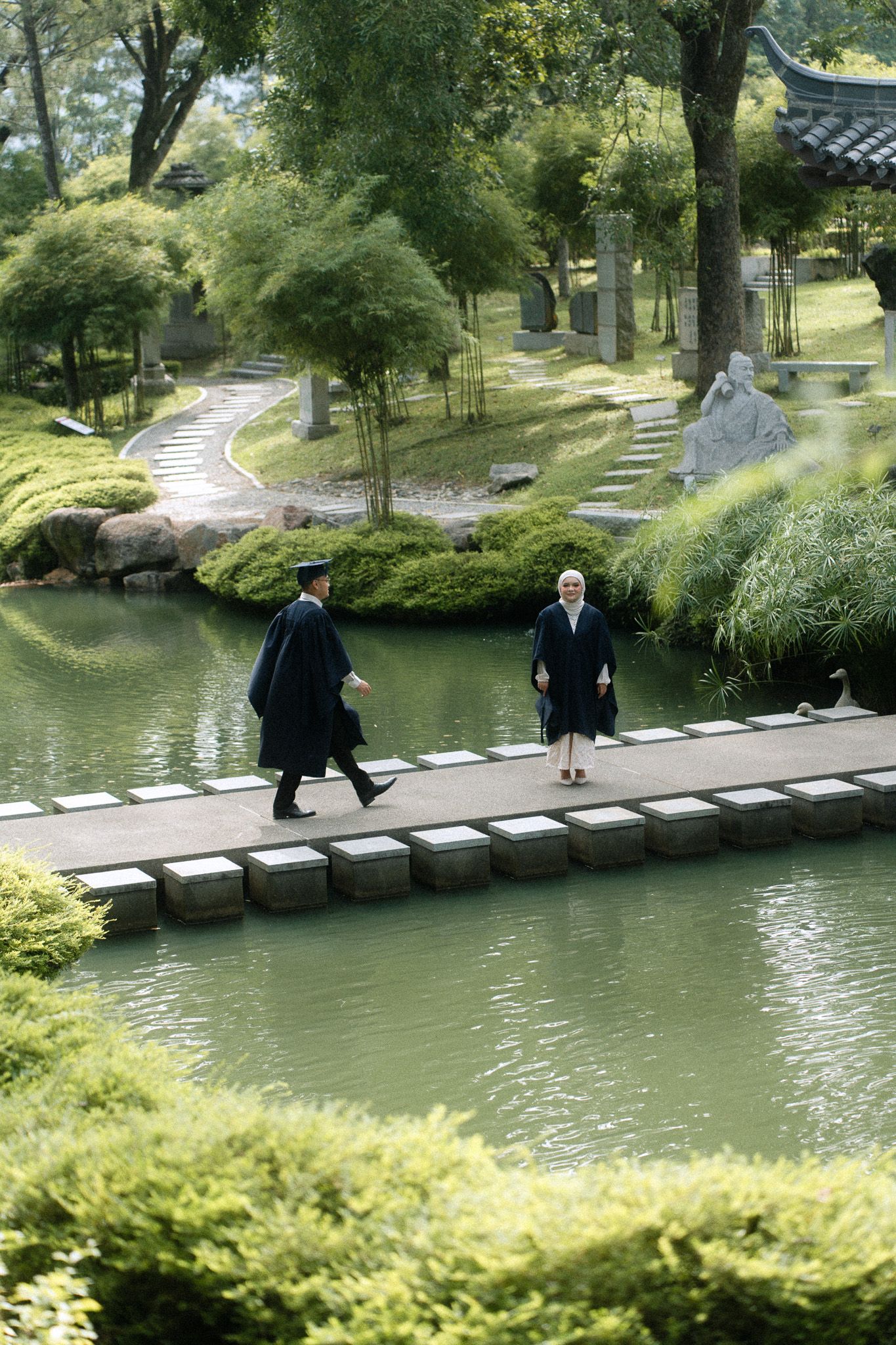 Two people in formal robes walk across a stone bridge over a serene pond in a lush Japanese garden.