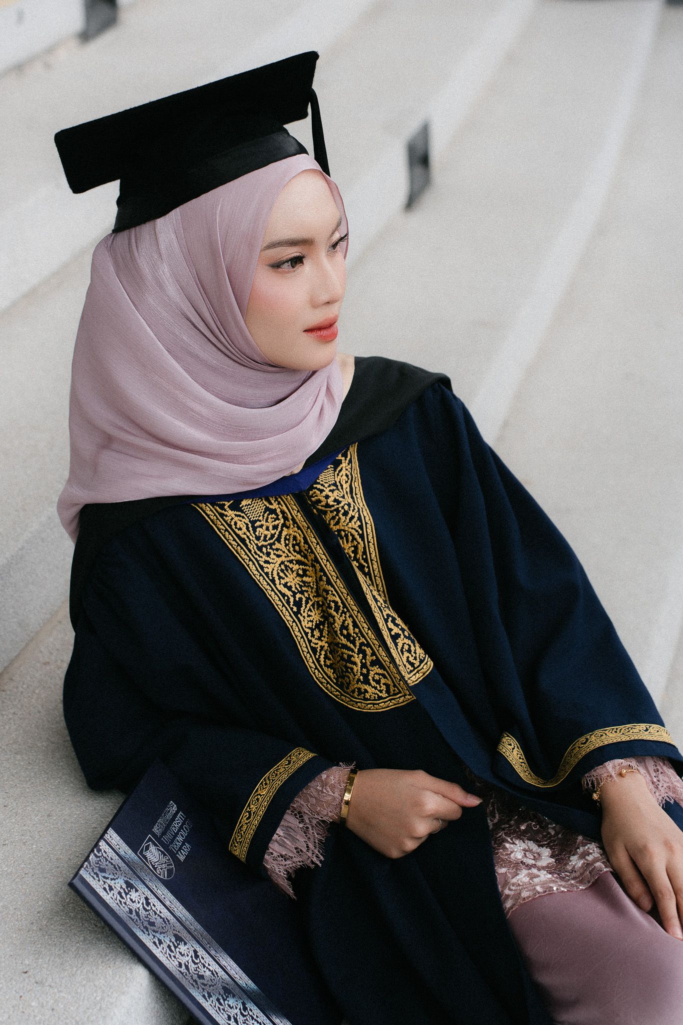 Young Muslim woman in graduation gown, hijab, and cap sitting on steps holding a diploma.