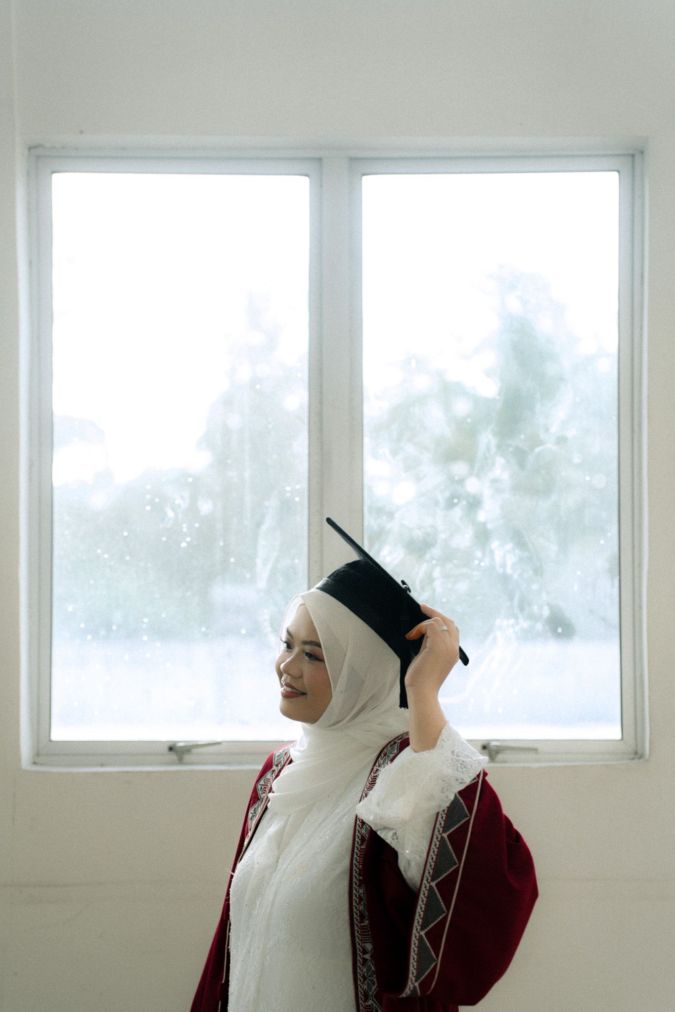 Smiling Muslim woman in hijab adjusting graduation cap by bright window in red academic gown.