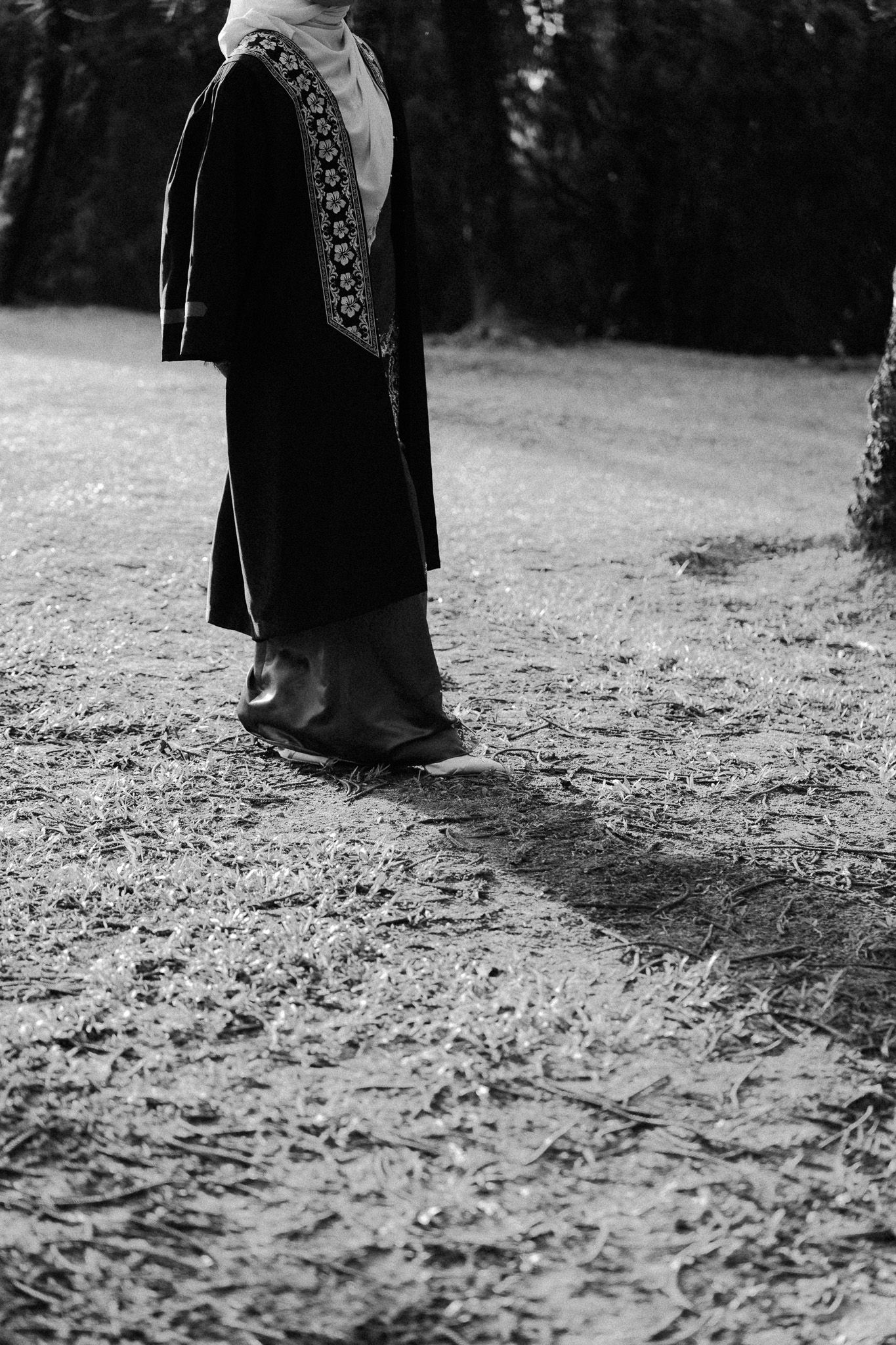 Black and white photo of a person in traditional dark robes and embroidered sash standing on a rural path.