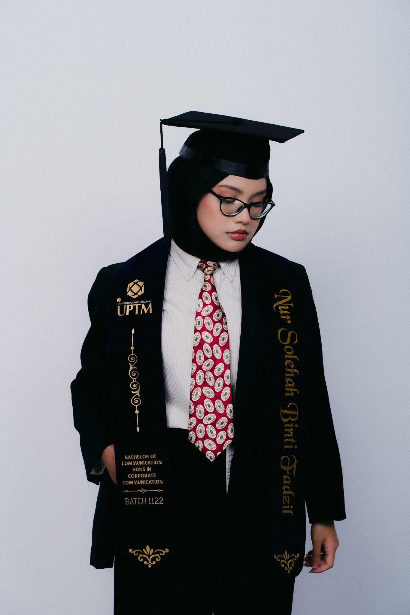 UPTM graduate in black gown and cap, wearing hijab, glasses, and red patterned tie, posing for graduation photo.