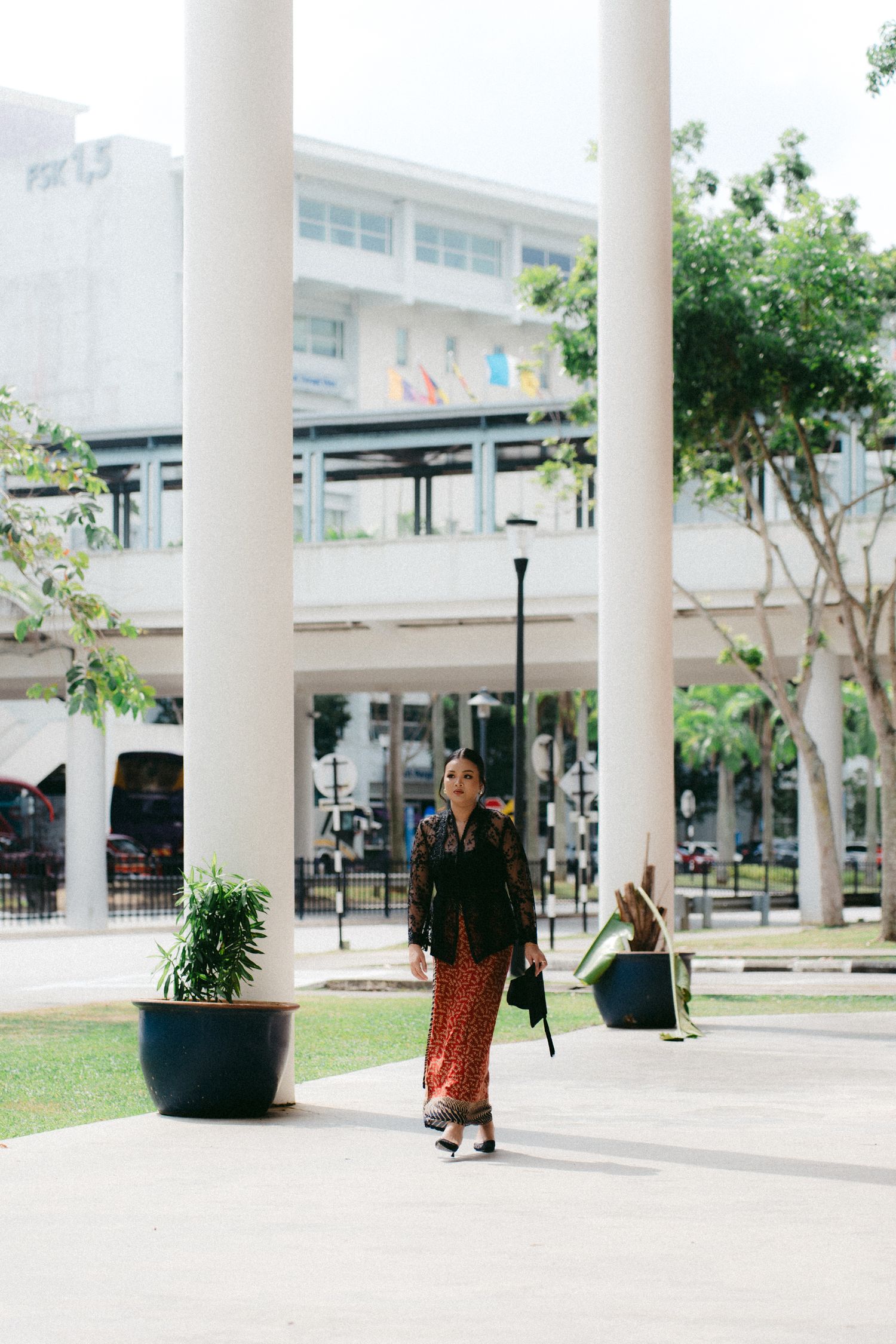 A person walks along a covered walkway with white columns and potted plants in a modern urban setting.