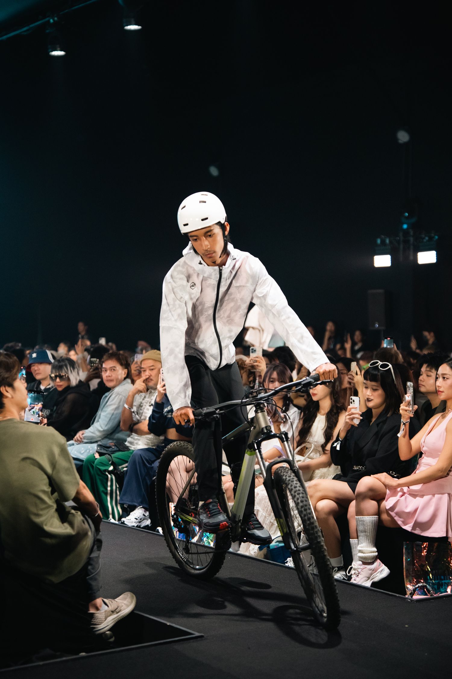 A performer rides a BMX bike on stage during a fashion show with audience members visible.