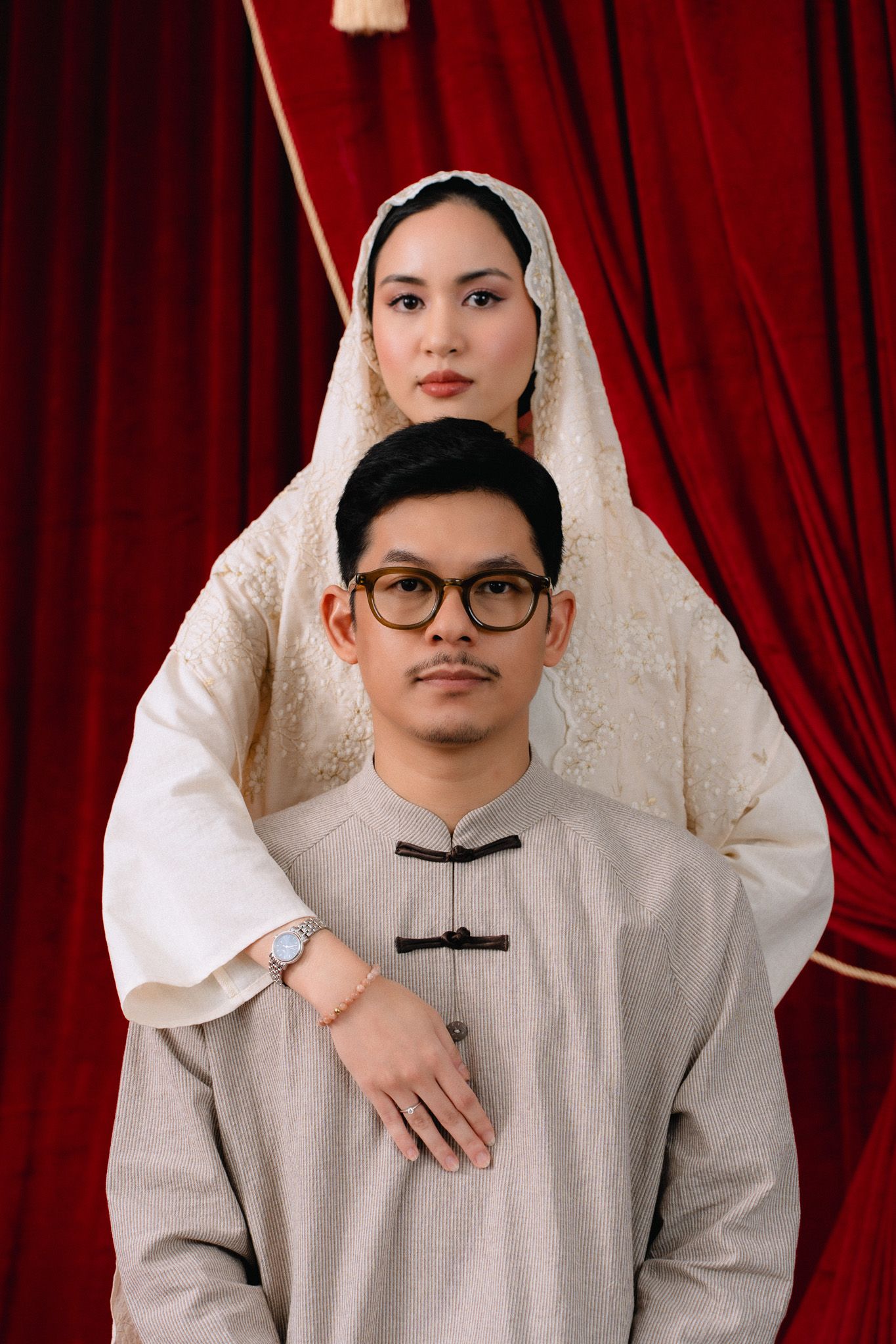A couple in traditional Muslim attire pose formally against red velvet curtains, man in gray baju kurung, woman in white hijab.