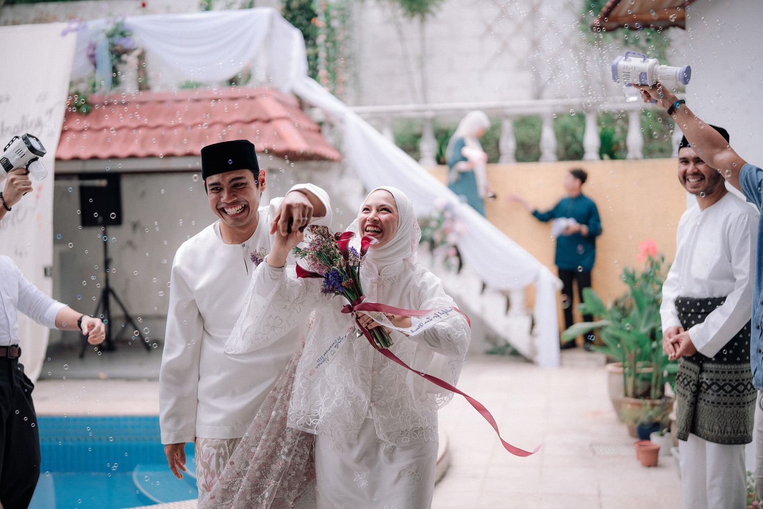 Smiling Muslim newlyweds in white traditional attire celebrating with guests at outdoor poolside wedding.