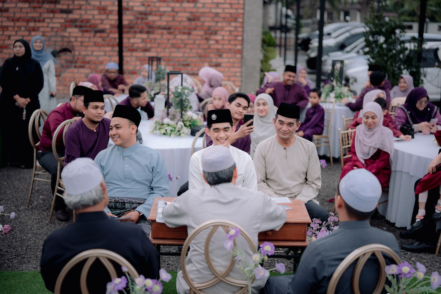 A traditional Malay wedding ceremony with guests in purple and gold baju melayu attire seated outdoors at decorated tables.
