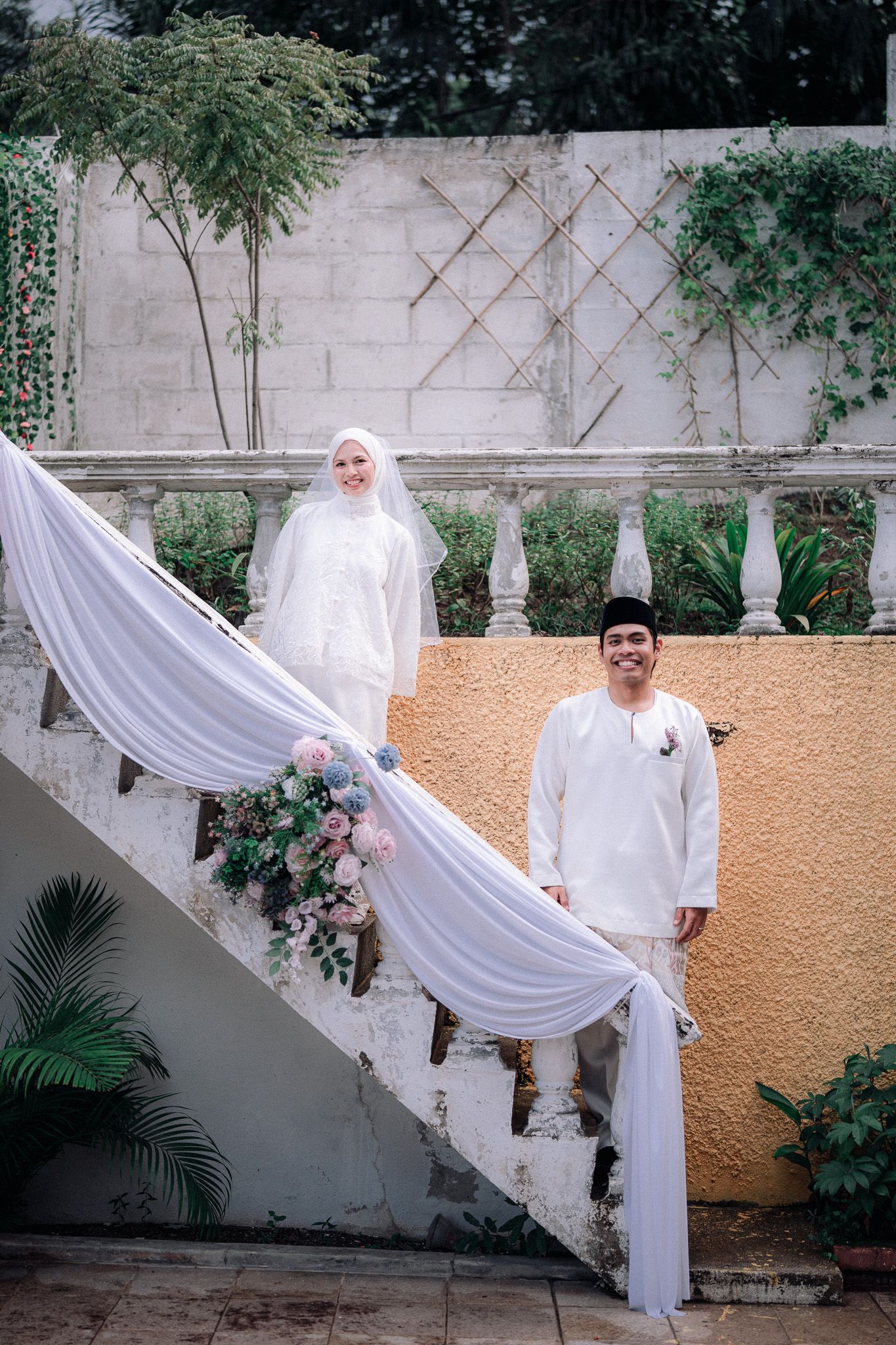 Muslim couple in white traditional attire posing on decorated stairs with floral arrangements at a wedding ceremony.
